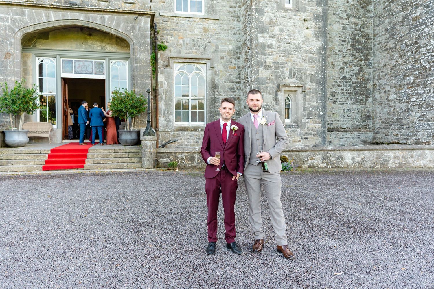 Wedding party members in burgundy and gray suits pose in front of a stone building entrance.