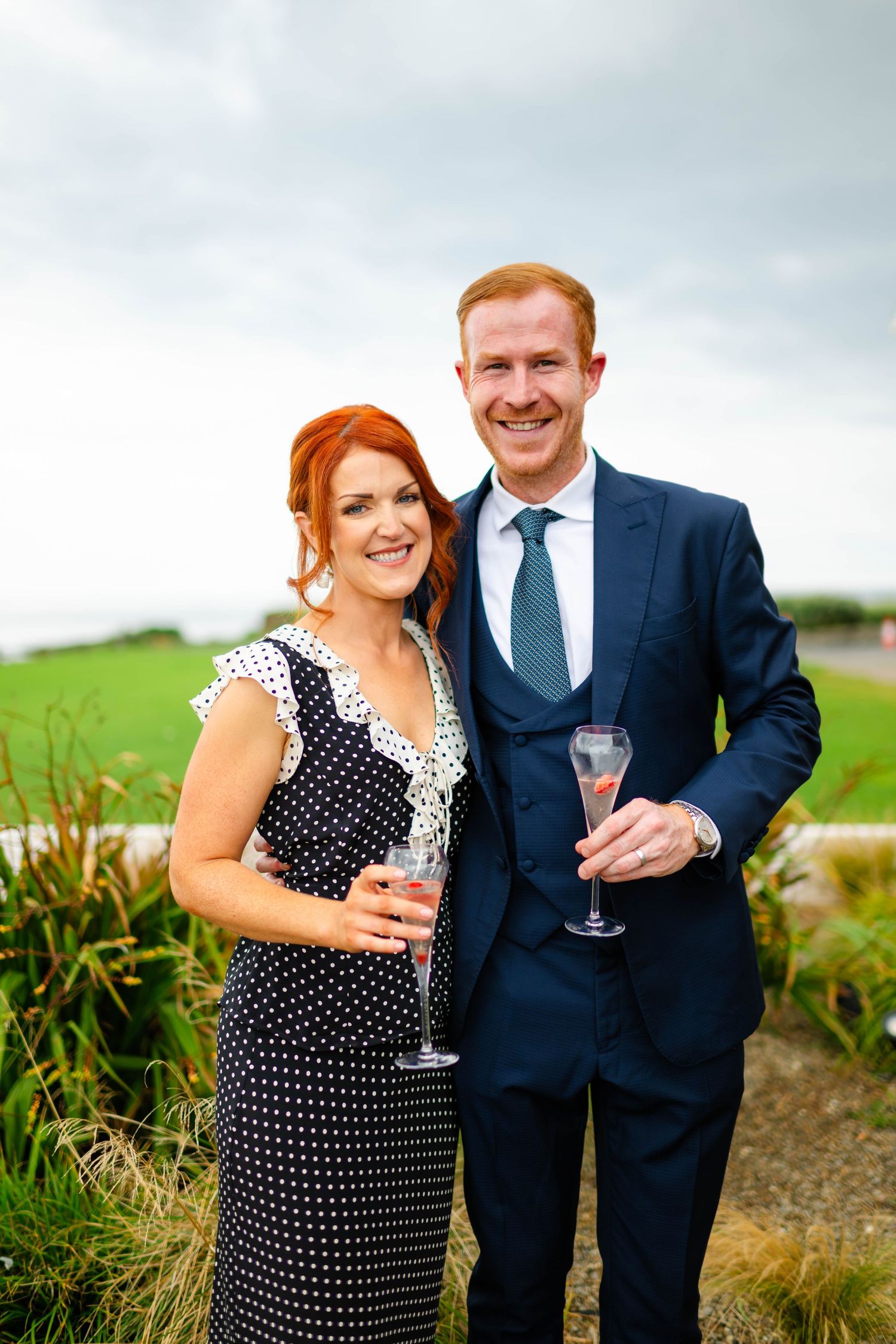 Couple in formal attire holding wine glasses pose together on grassy hillside during outdoor celebration.