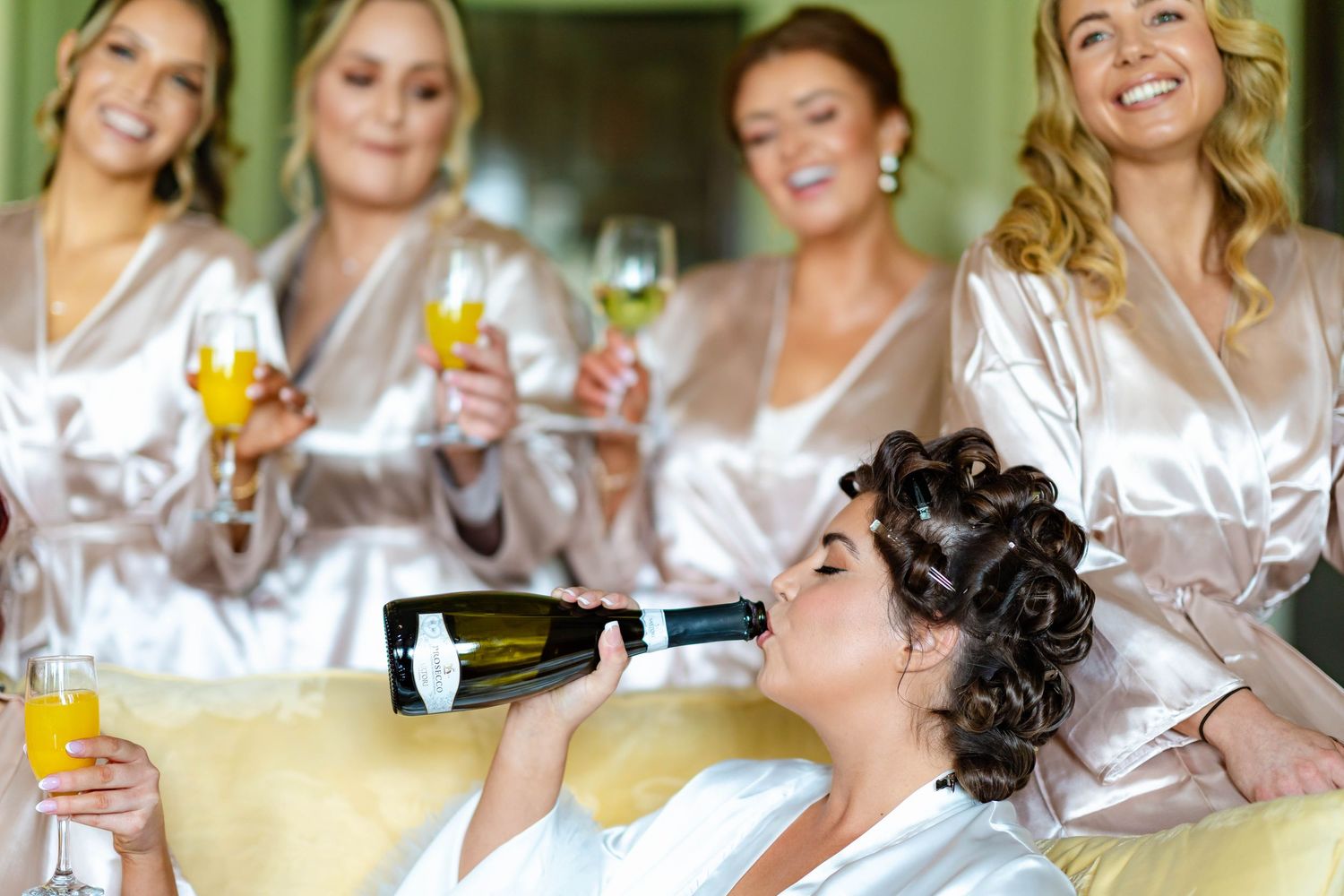 Wedding party members in matching white robes share laughs and drinks while getting ready for the celebration.