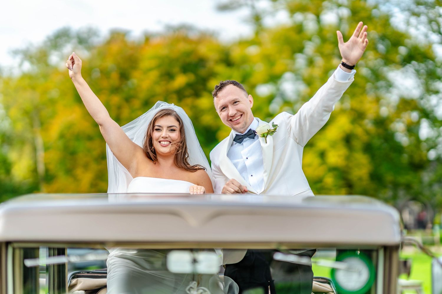 Happy couple celebrates with arms raised while riding in a golf cart at their wedding ceremony.