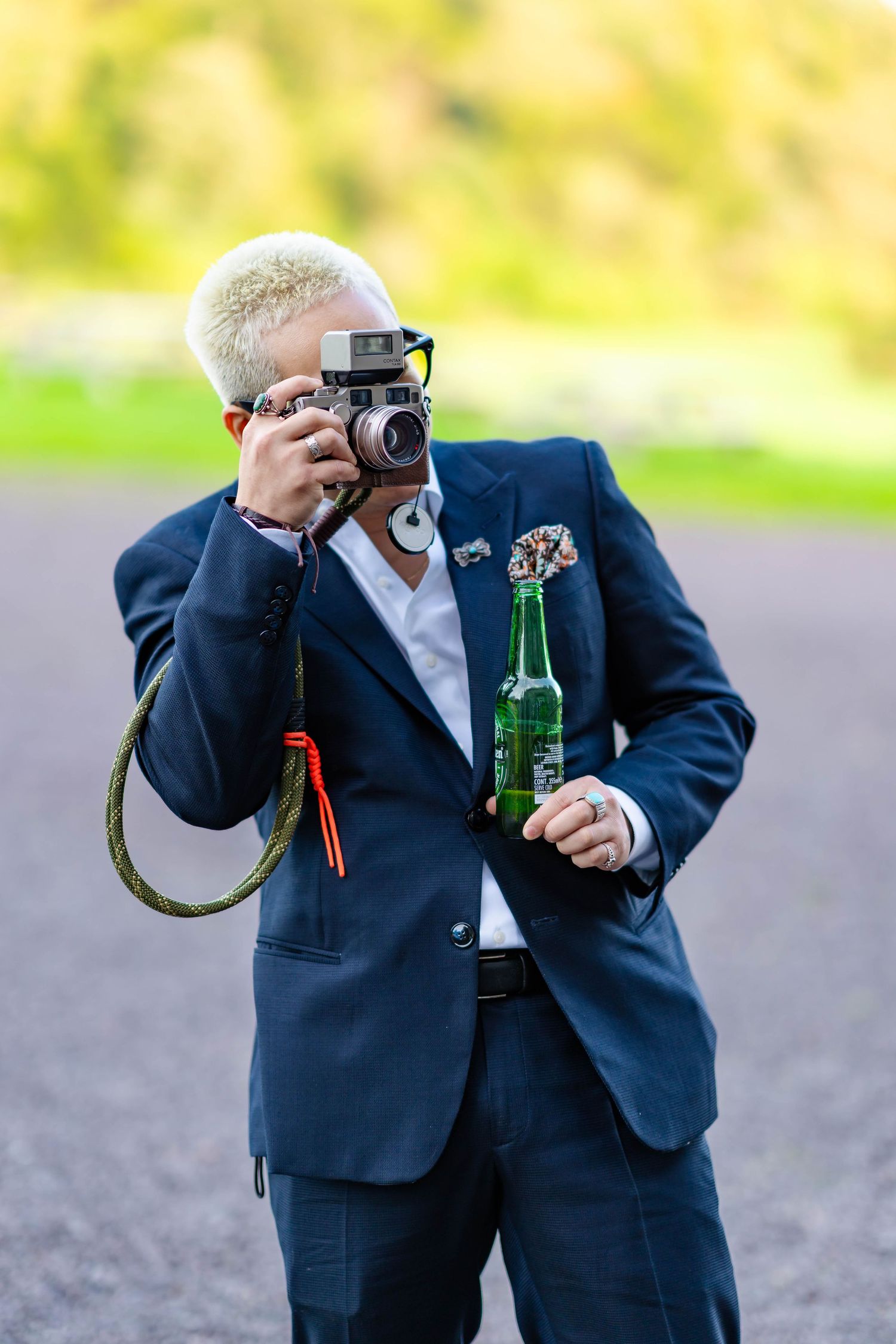 A photographer in a navy suit jacket holds up a camera while drinking from a green glass bottle.