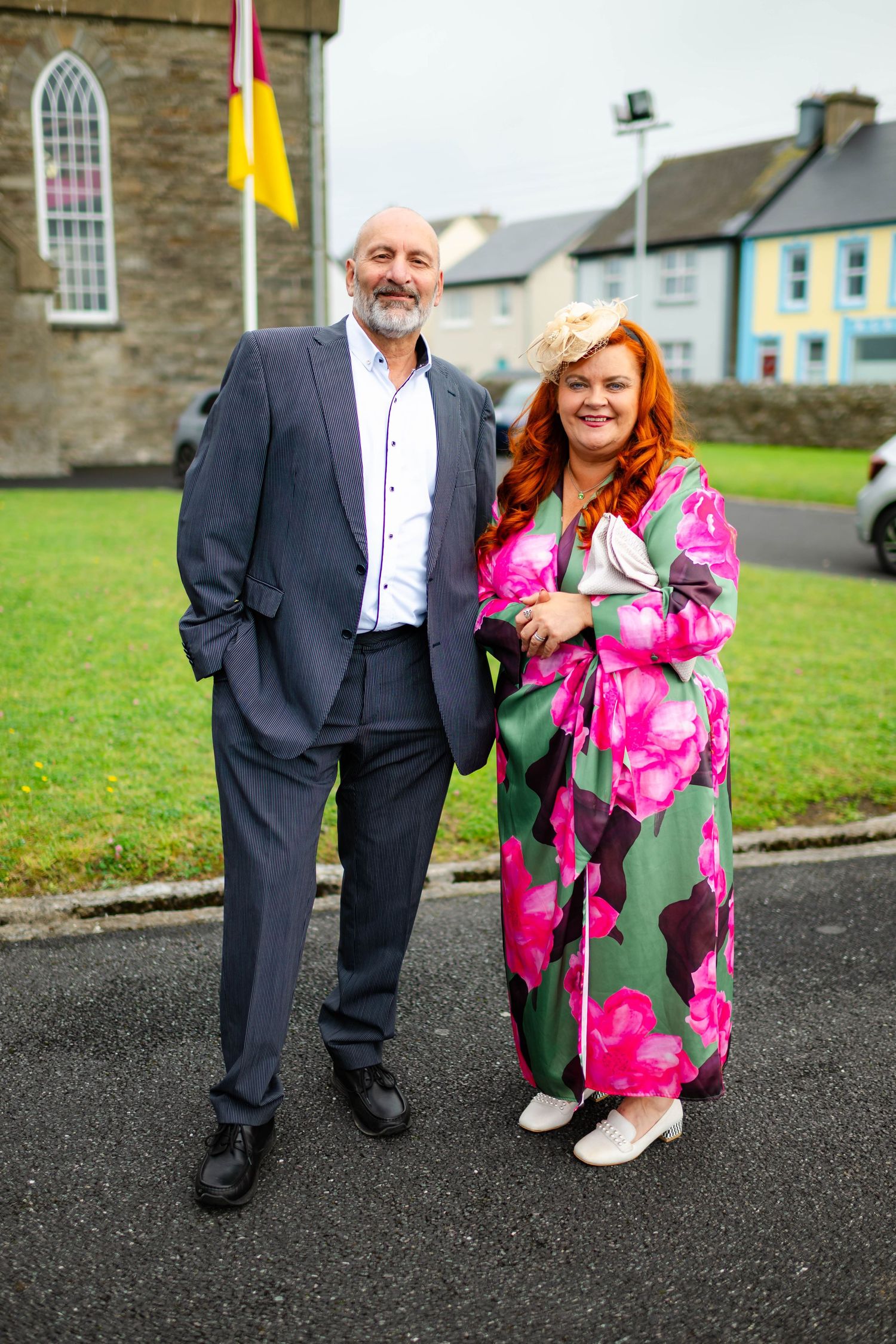 Guests in formal attire posing together on asphalt with colorful buildings in background.