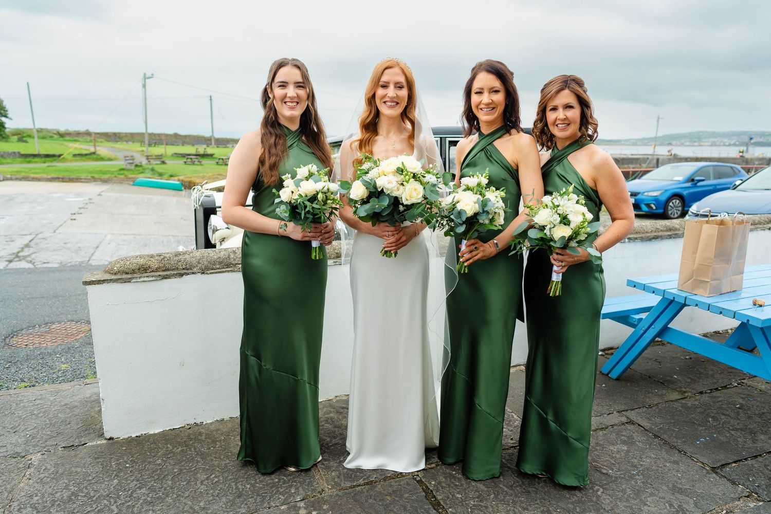 Bridal party in emerald green dresses poses by waterfront with bouquets.