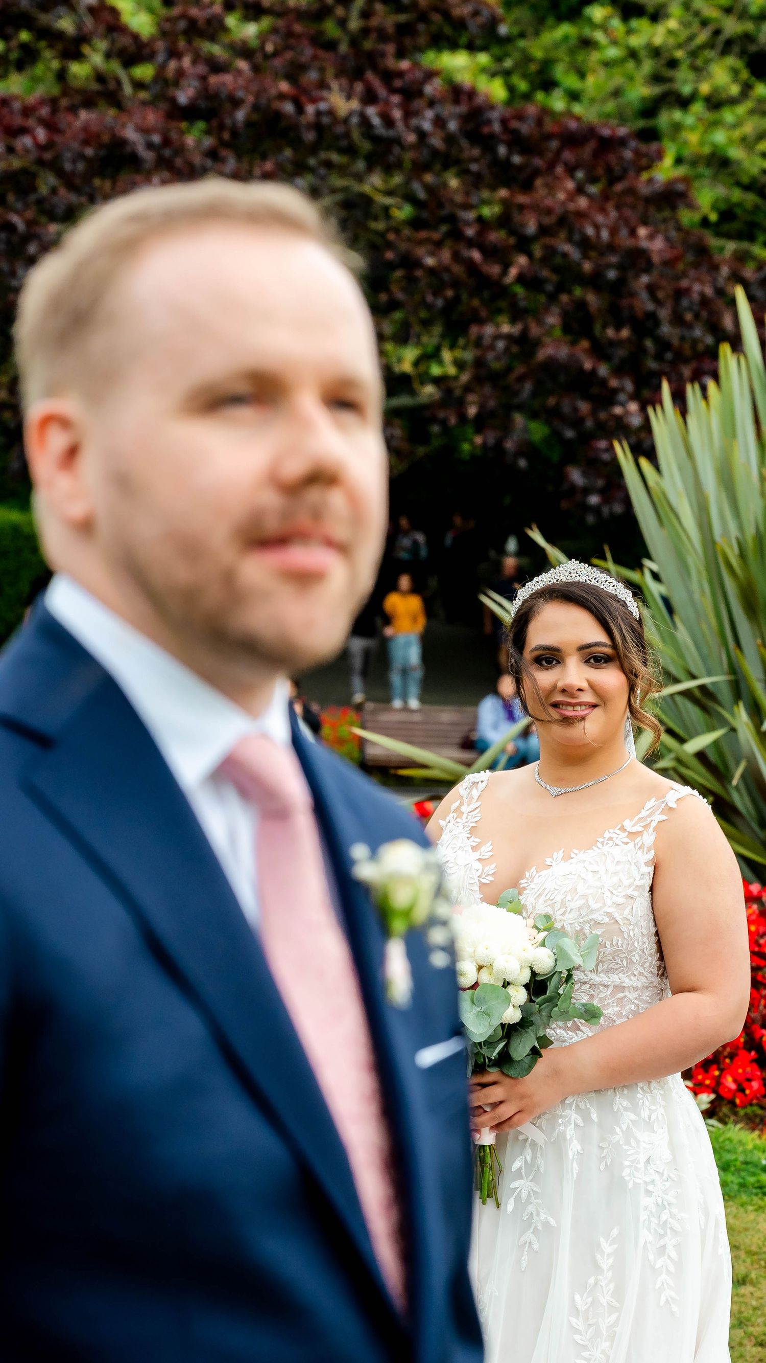 A couple poses in a garden with red flowers and palm plants in the background.