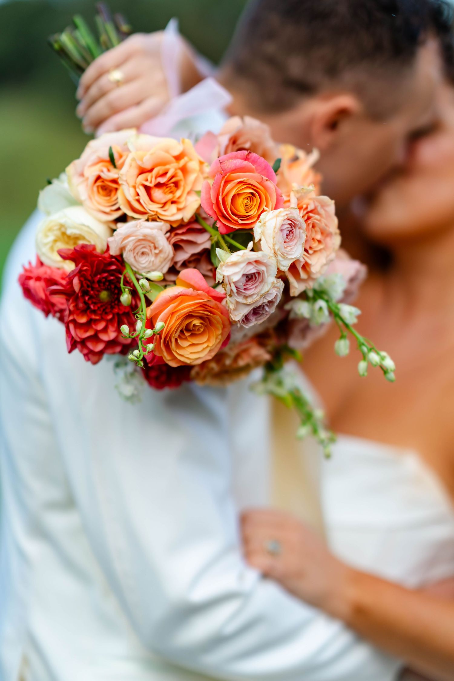 A wedding bouquet featuring peach, coral and red roses with delicate greenery against a white dress background.