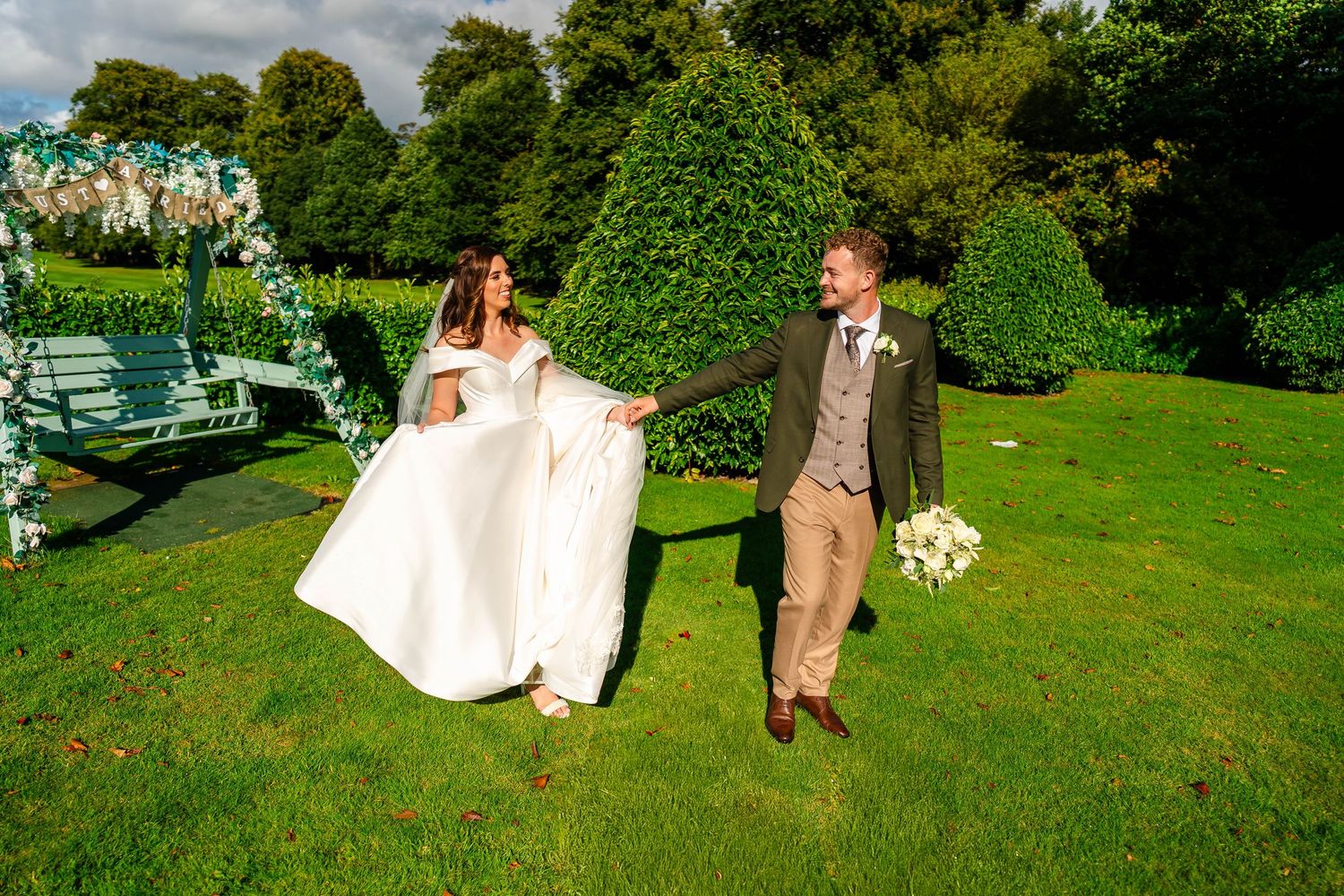 Wedding party poses for photos on manicured lawn with white gazebo and greenery in background.