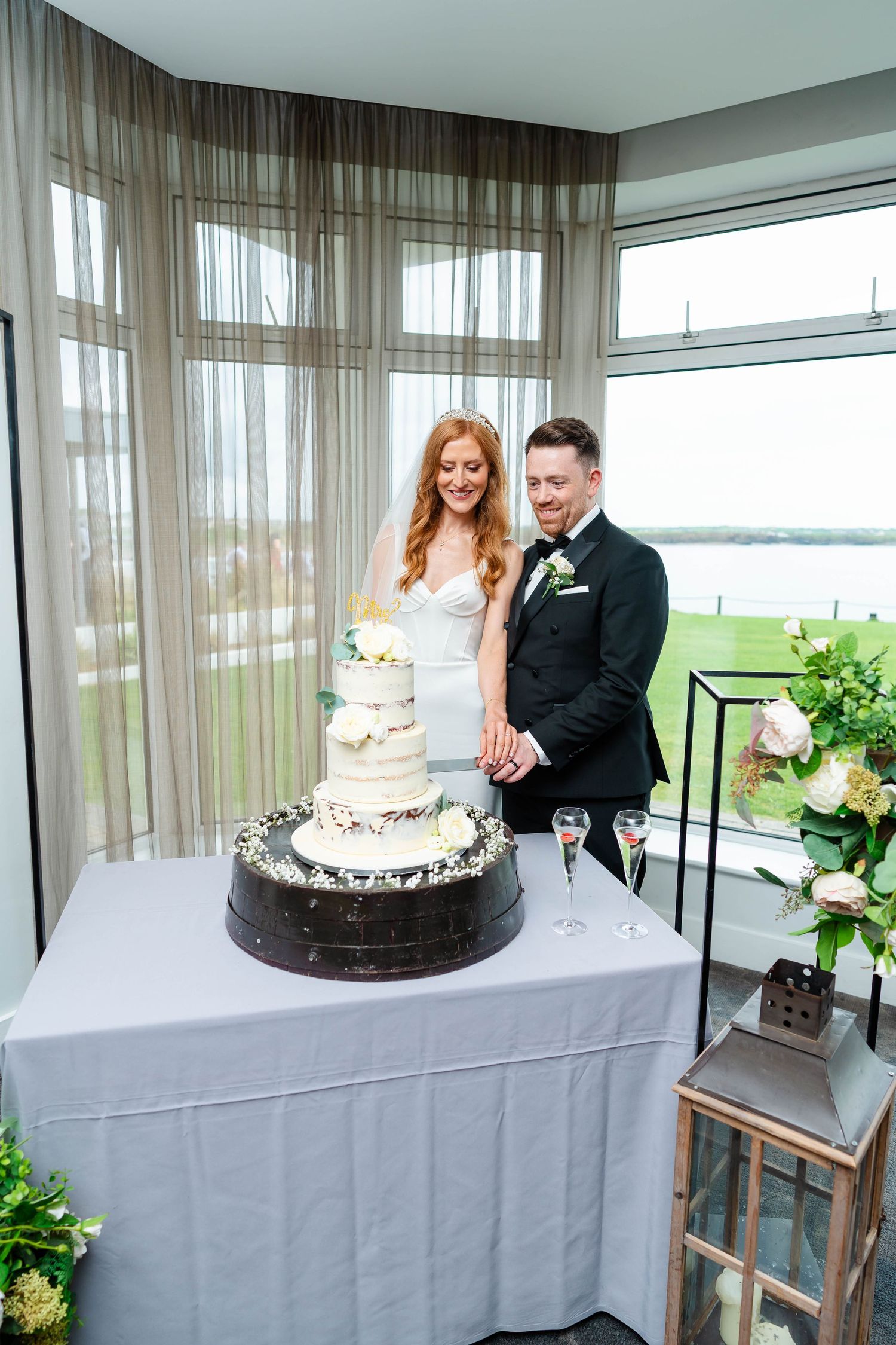 A wedding cake display table with white linens and sheer curtains overlooking a waterfront view.