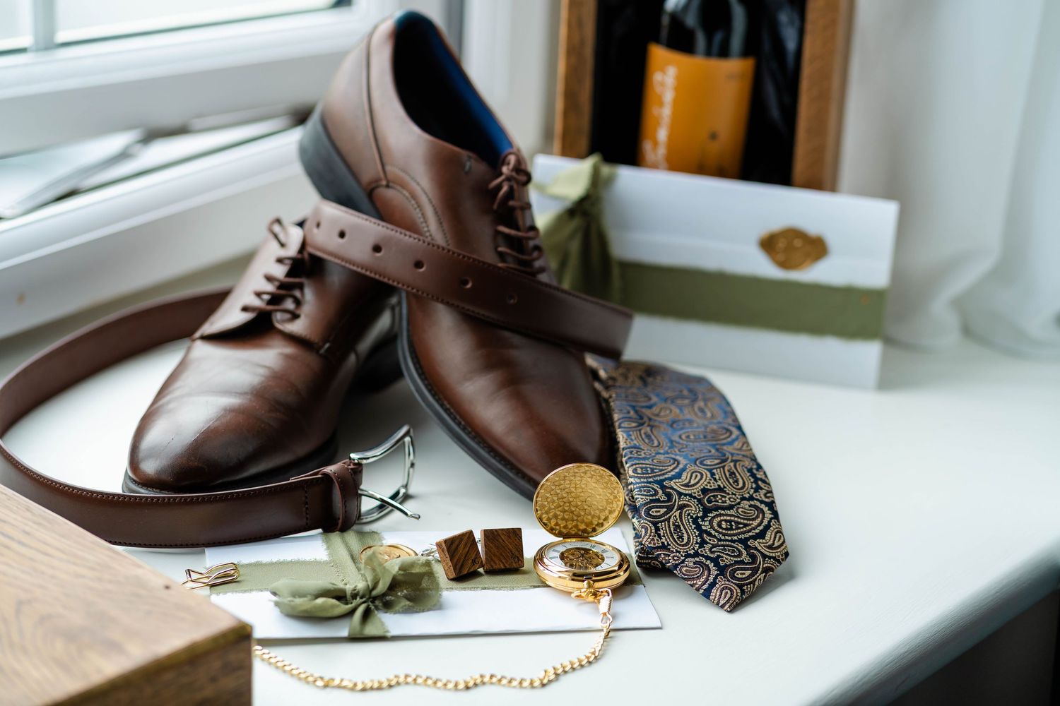 Wedding details featuring brown leather dress shoes and accessories on white windowsill.