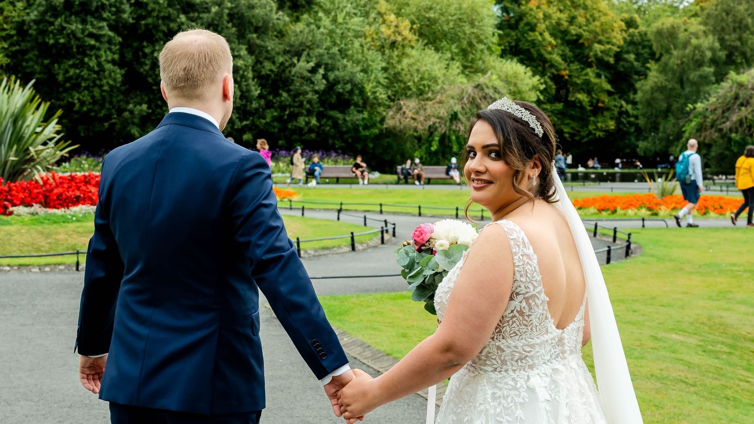 Wedding couple walking through a scenic park with vibrant greenery and red flowers in the background.