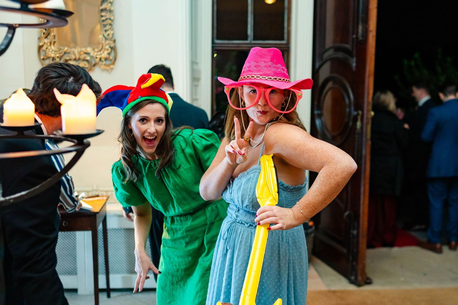 Two people in colorful outfits, one in green dress and one in blue with a pink cowboy hat, pose playfully at an event.