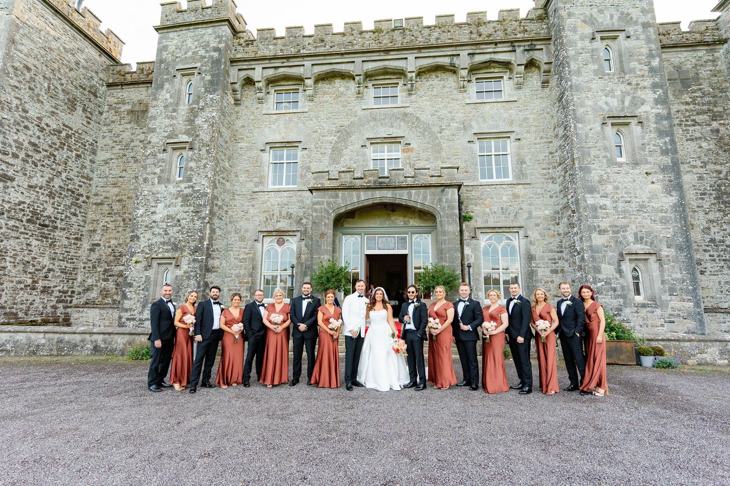 Wedding party poses in front of historic stone castle walls during outdoor celebration.