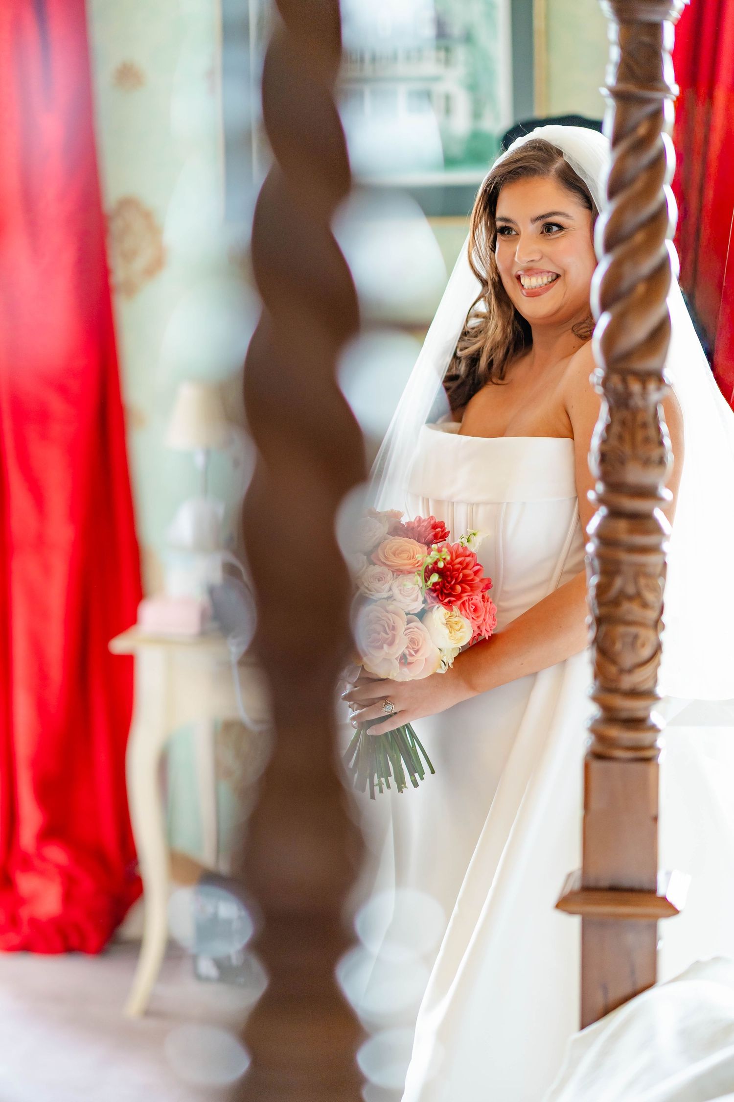 A bride in a white wedding dress holds a coral-colored bouquet while standing between decorative wooden pillars.