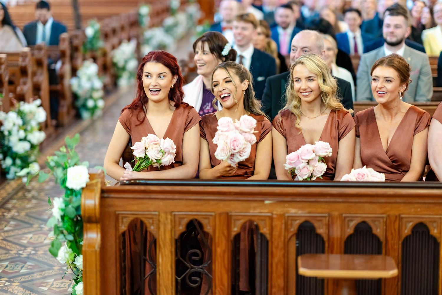 Wedding party laughs together while seated on wooden church pews decorated with floral arrangements.