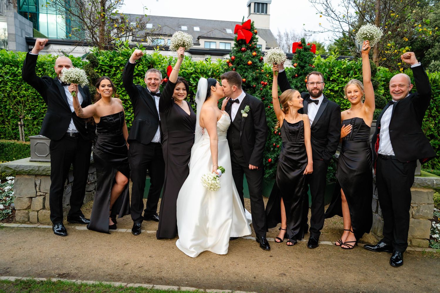 Wedding party celebrates with raised arms and joyful expressions in outdoor garden setting.