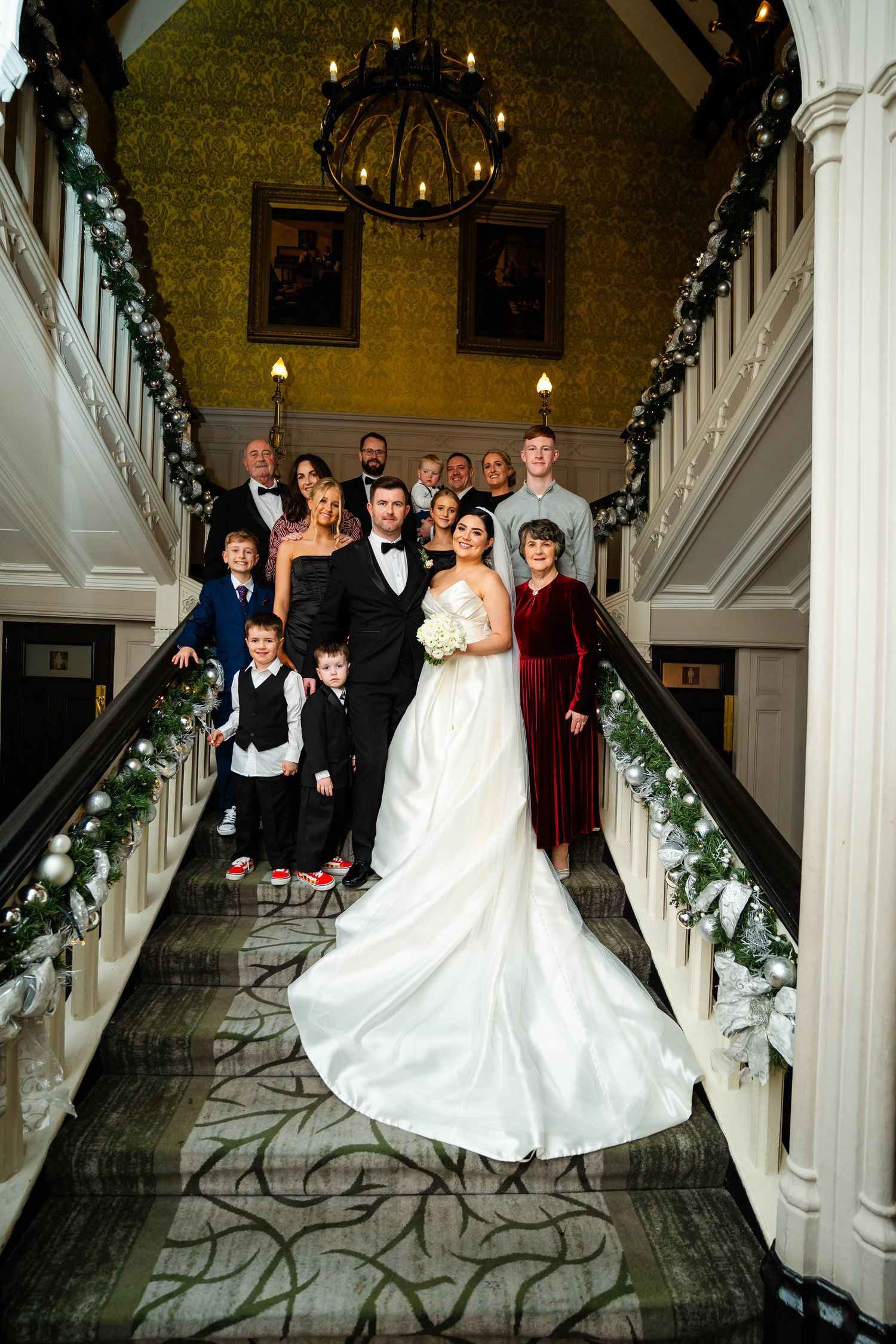 Wedding party poses on grand staircase decorated with floral garlands and white architectural details.