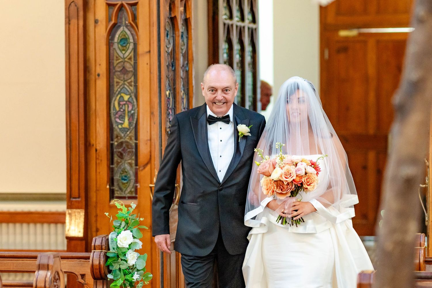 A wedding ceremony in a church with a bride in white veil holding peach roses and someone in a black tuxedo.
