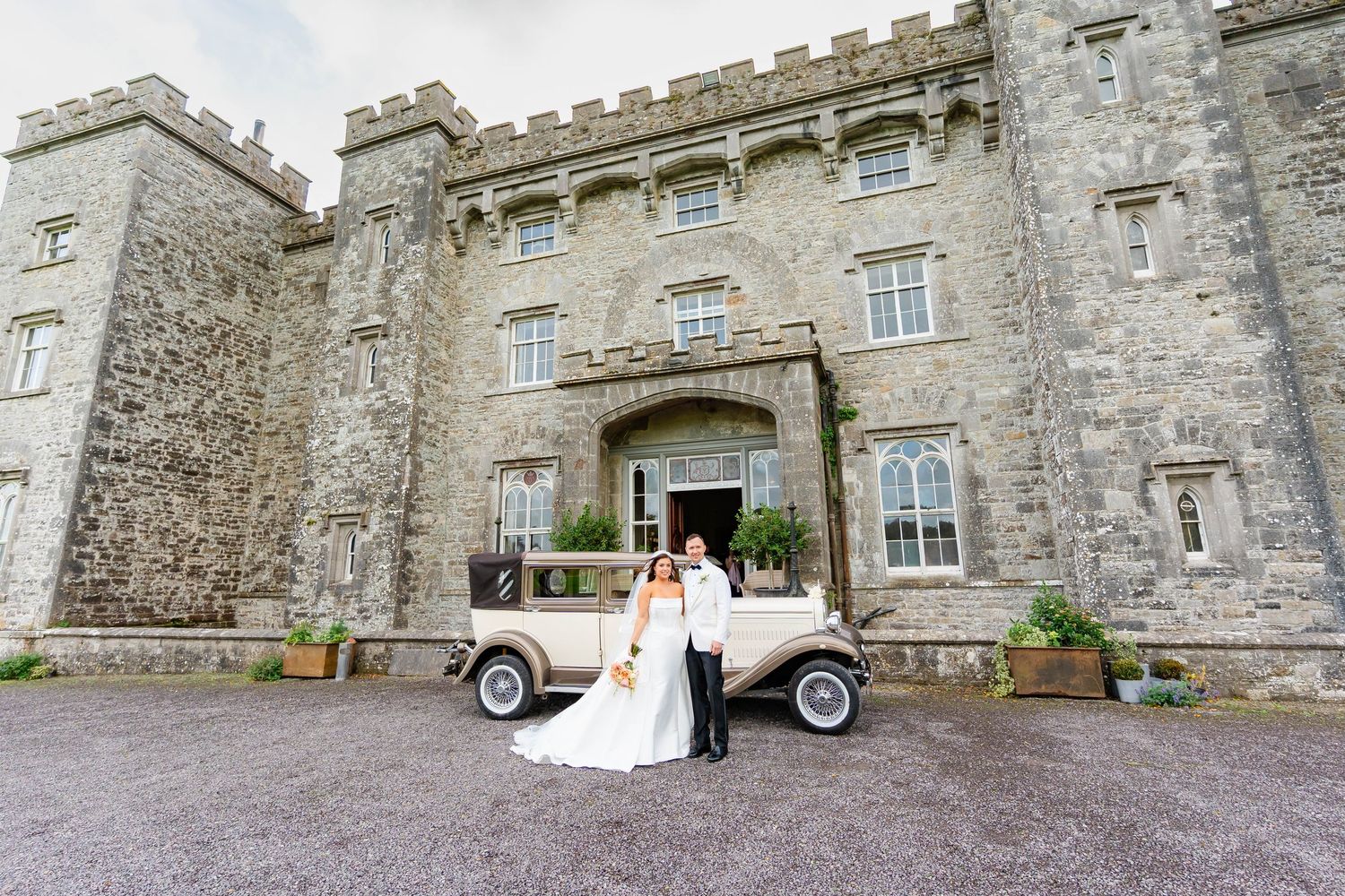 A classic vintage car parked in front of a grand stone castle entrance with wedding decorations.