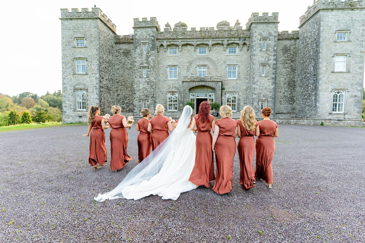 Wedding party in coral dresses poses for photos in front of a grand stone castle.