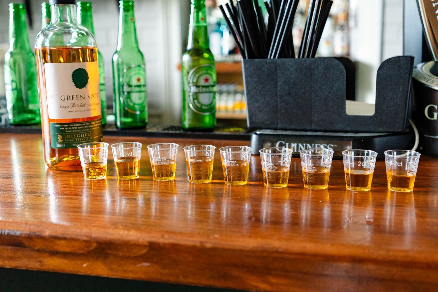 A wooden bar counter displays various liquor bottles and glasses in a row with green lighting in the background.