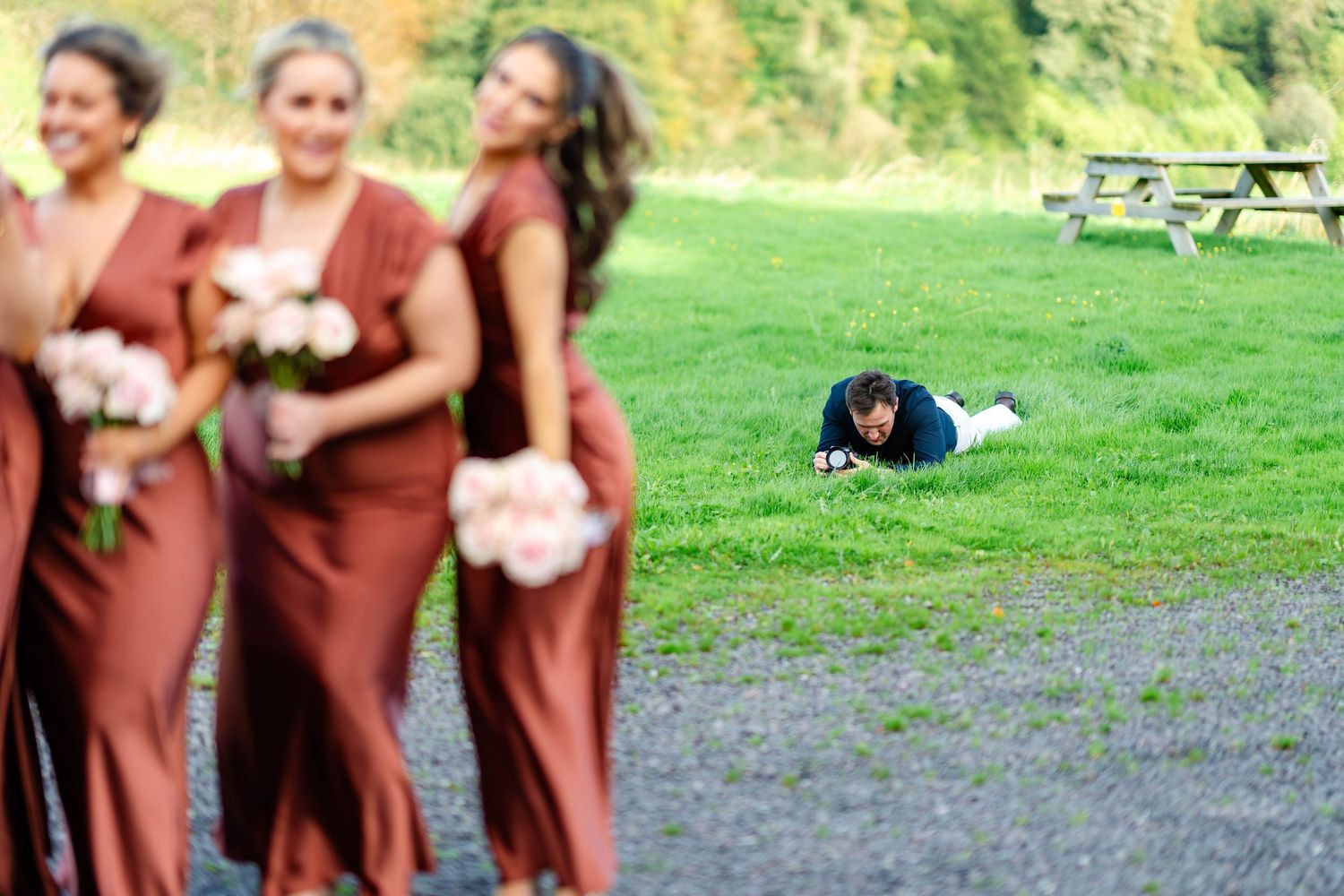 A wedding party in burgundy dresses poses on a grassy lawn near a picnic table on a sunny day.