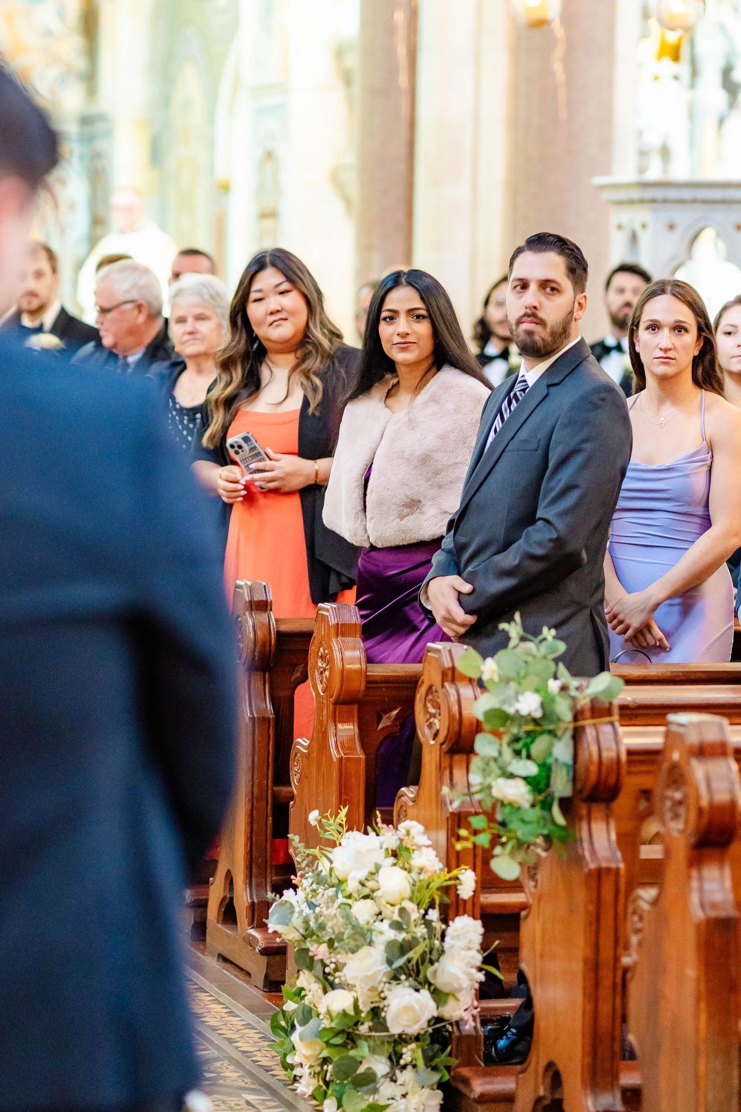 Wedding guests seated in wooden church pews decorated with white floral arrangements during ceremony.