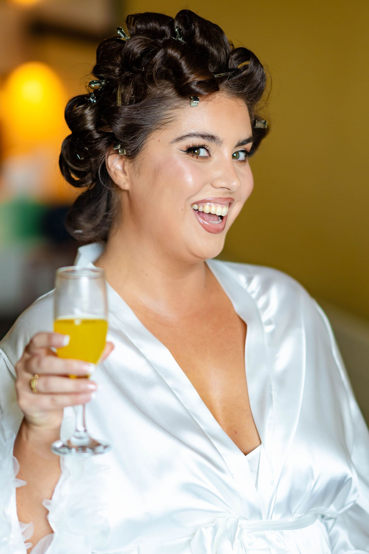 Person wearing white silk robe holds champagne glass while smiling with styled dark curls during celebration.