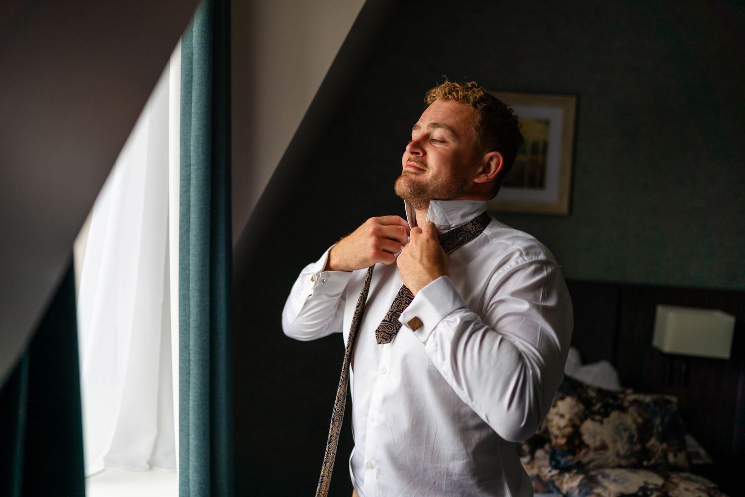 Person adjusting tie while looking out window in moody lighting.