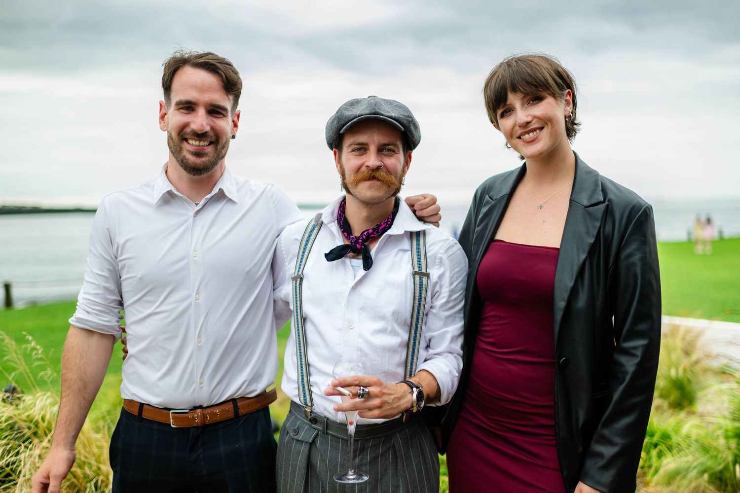Group of wedding guests pose together on grassy lawn by the coast.