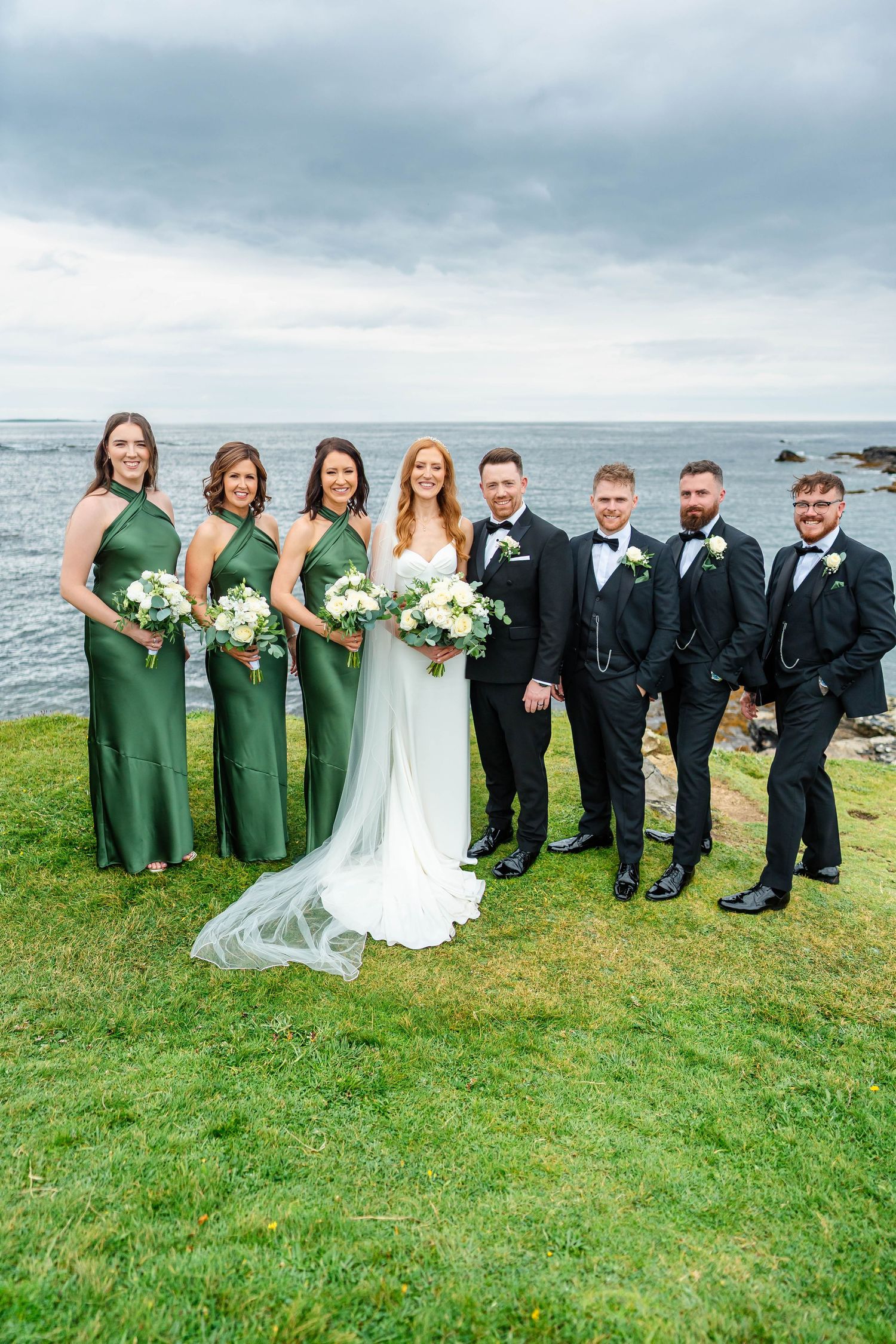 Wedding party members in emerald green dresses and black tuxedos line up for formal photos by the sea.