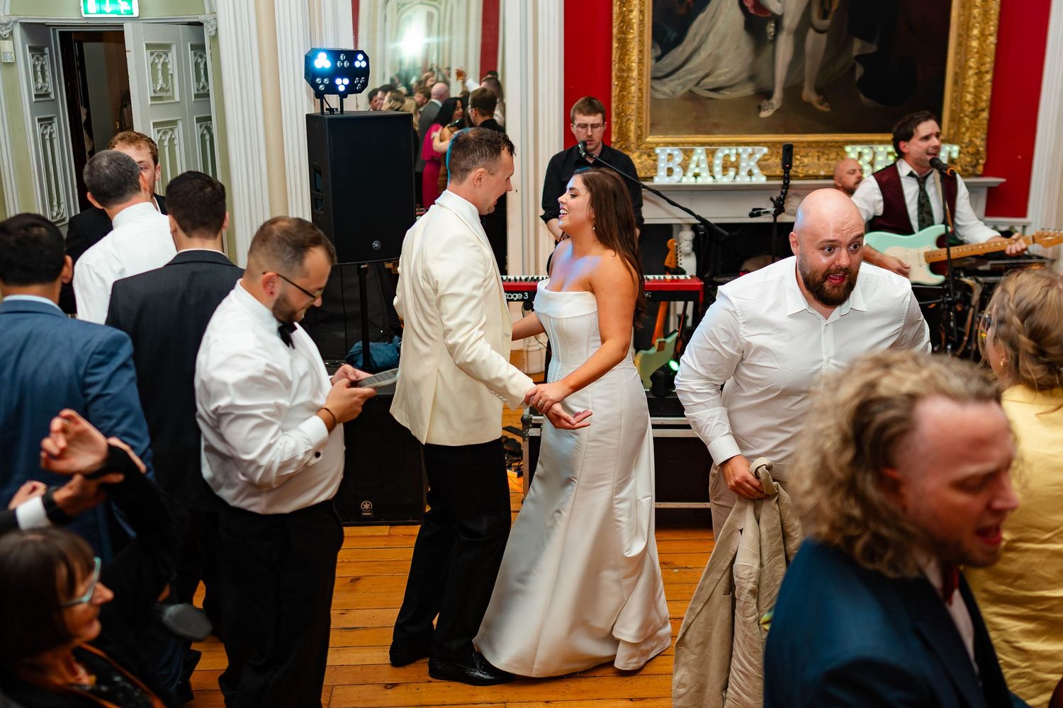Wedding guests dance and celebrate together on a wooden dance floor at an indoor reception venue.