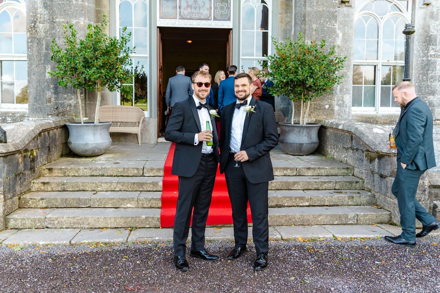 Two people in tuxedos stand together on stone steps outside an elegant building with potted plants.