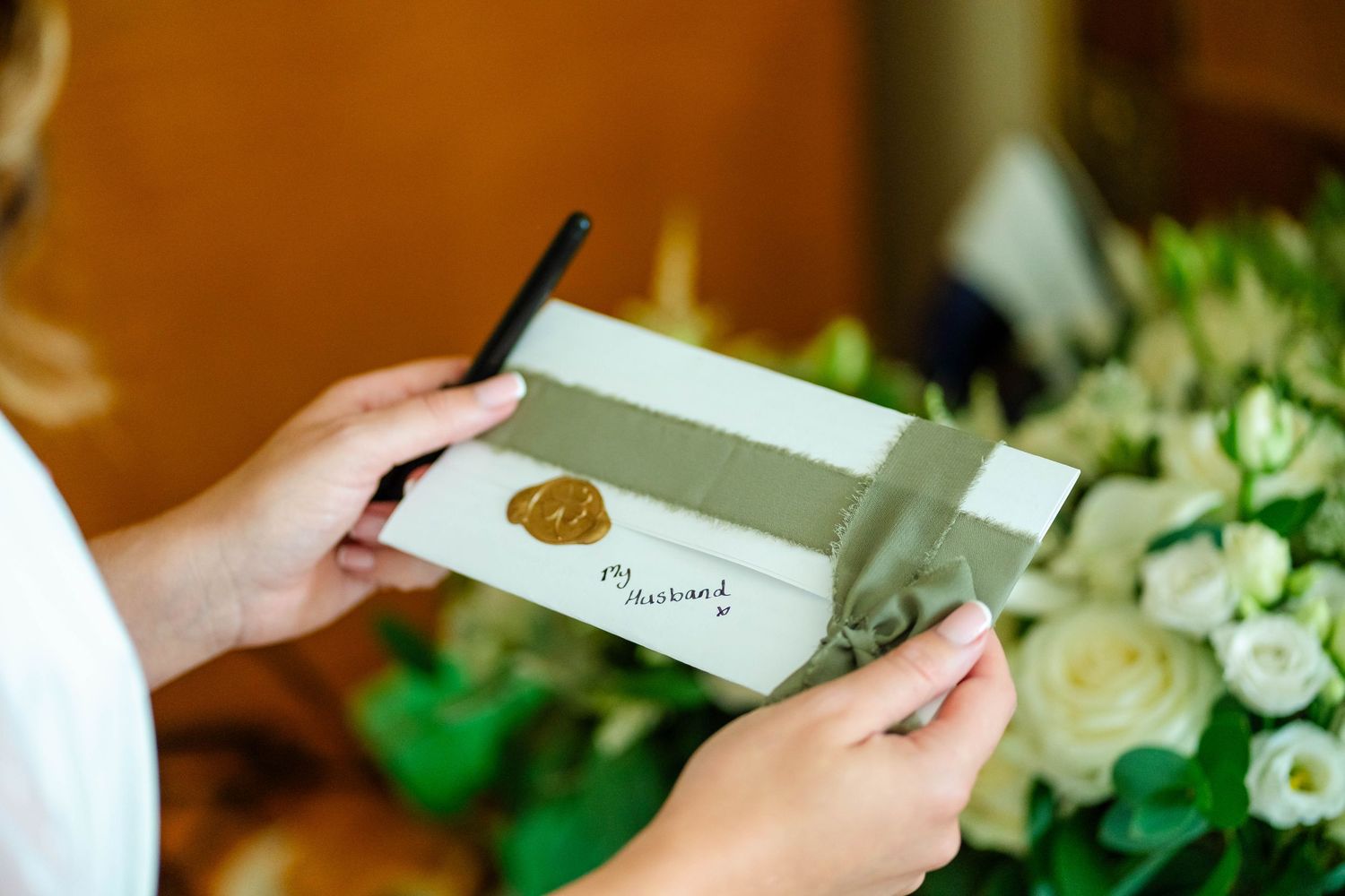 Hands holding wedding invitation cards with white roses and greenery in background.