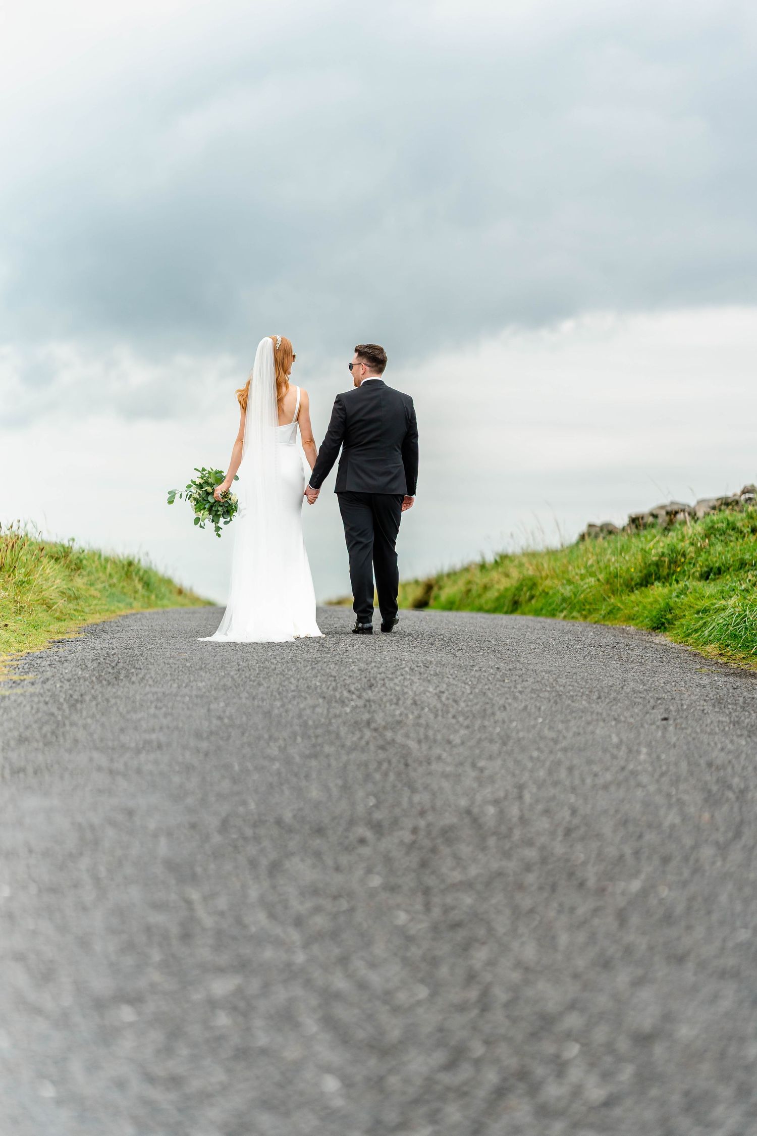 Silhouetted wedding couple walking up paved hill against cloudy sky.