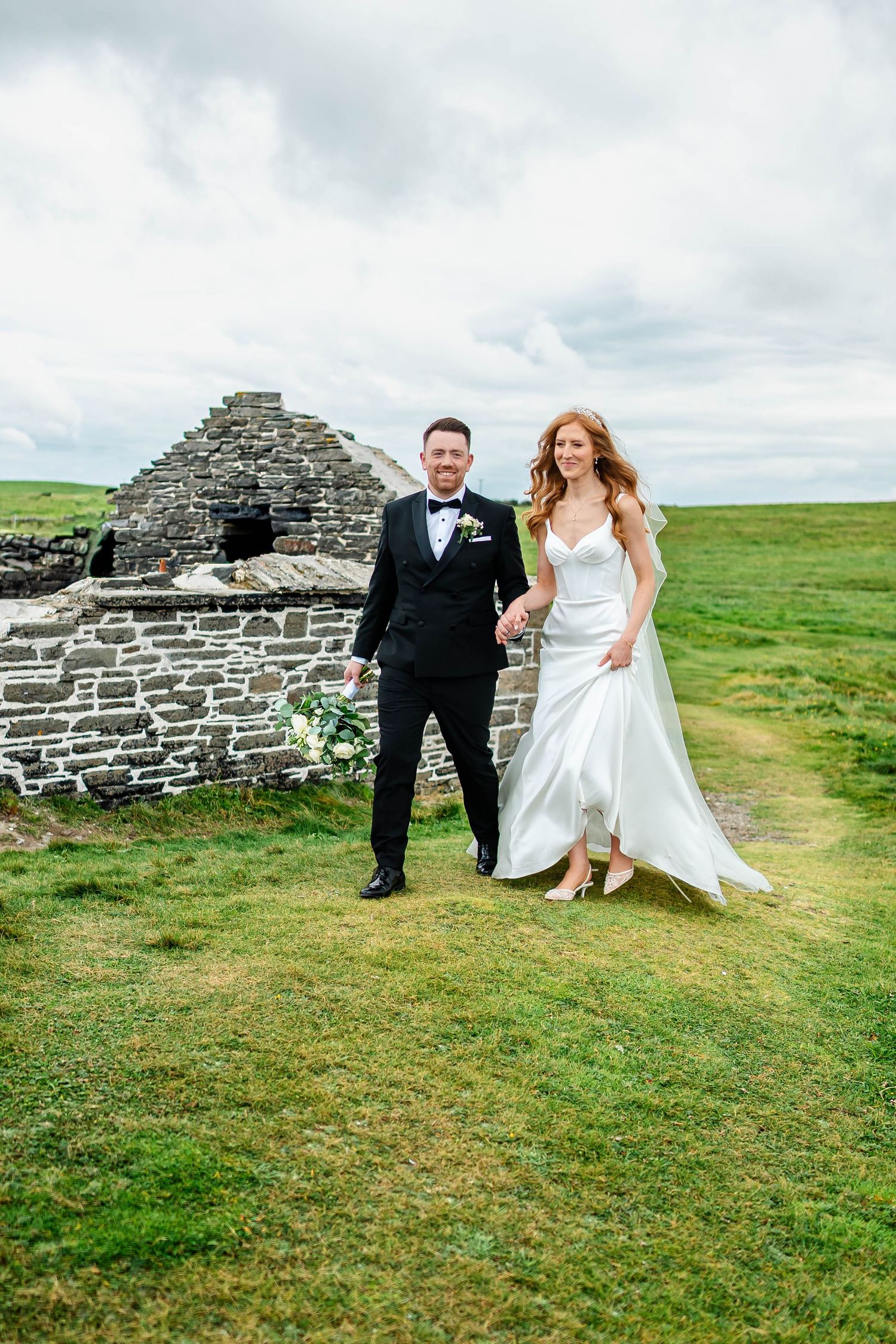 Wedding couple poses playfully near an ancient stone structure in the Irish countryside.