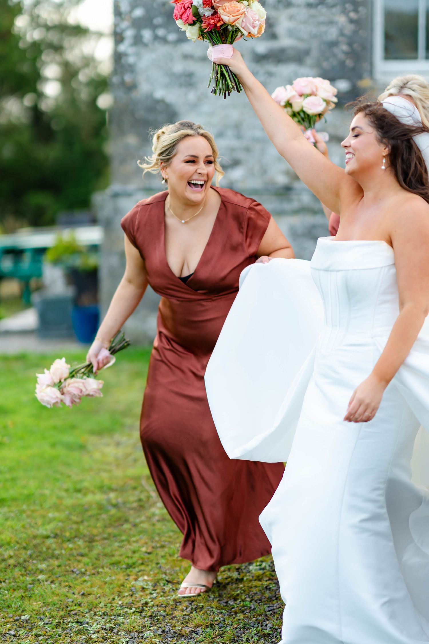 Two wedding attendees share a joyful moment outdoors, one in burgundy dress and one in white gown holding bouquets.