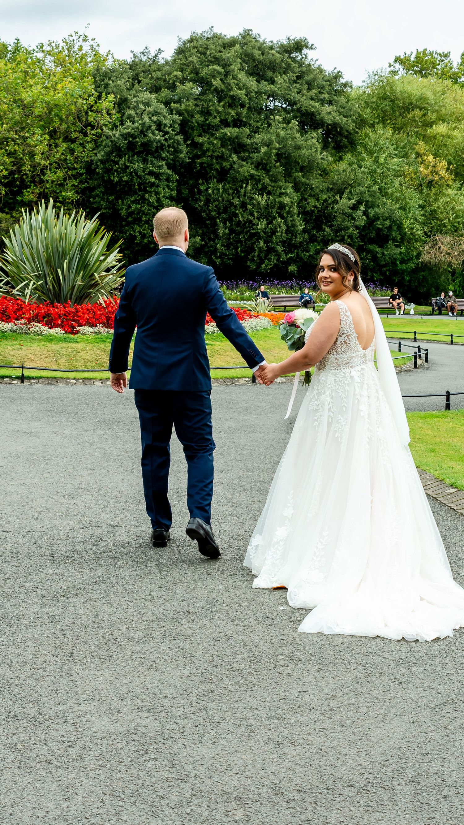 Newlyweds strolling along a park pathway with beautiful landscaping and red flower beds.