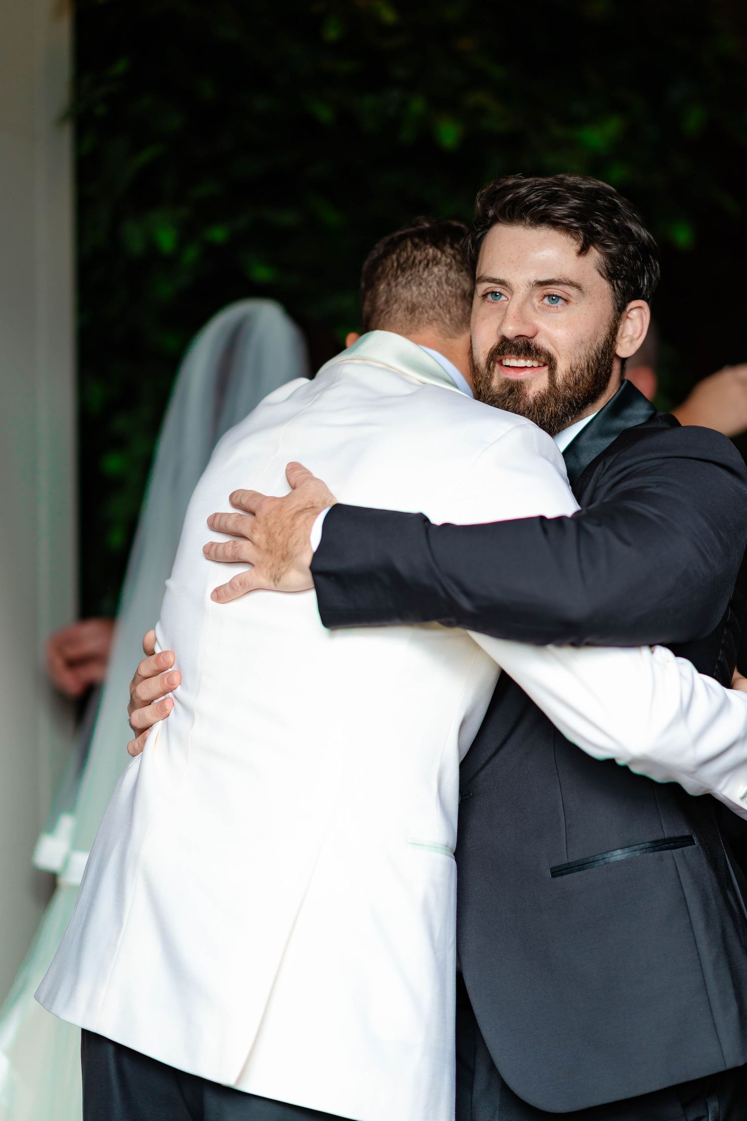 Two men in formal suits share a warm embrace at a wedding celebration.