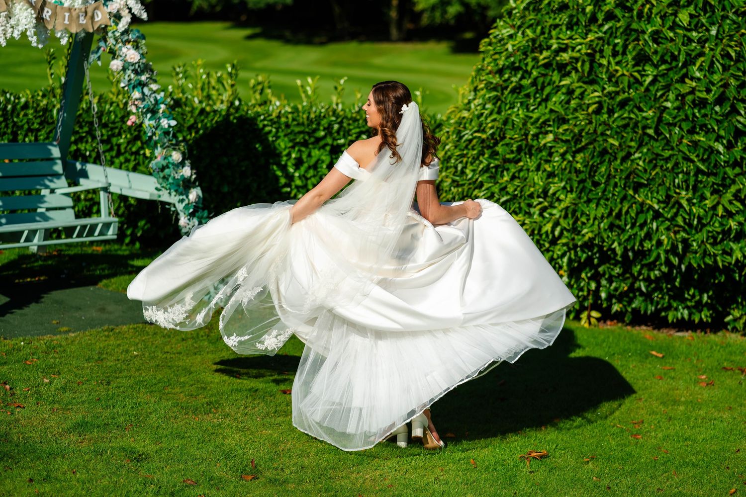 Bride's dress flows gracefully across green lawn during outdoor wedding photoshoot.