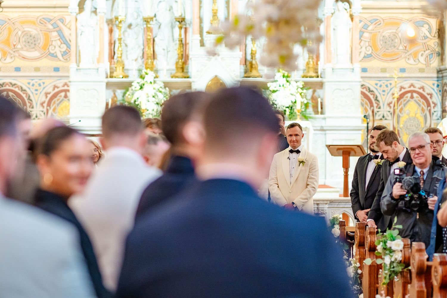 Wedding guests gather in an ornate church interior with decorative columns and stained glass windows during a ceremony.