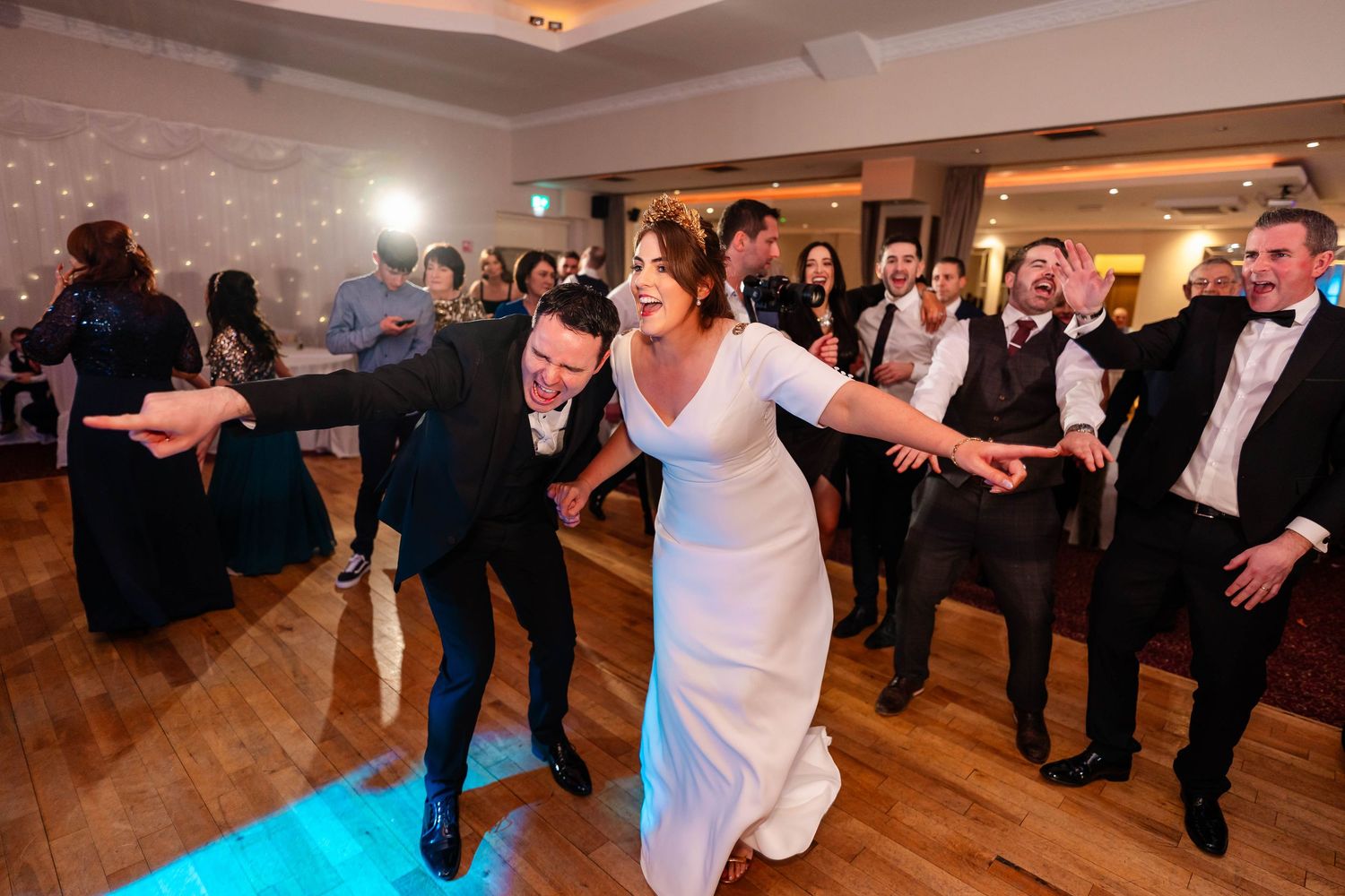 Wedding party dancing and celebrating on a wooden dance floor at an indoor venue.
