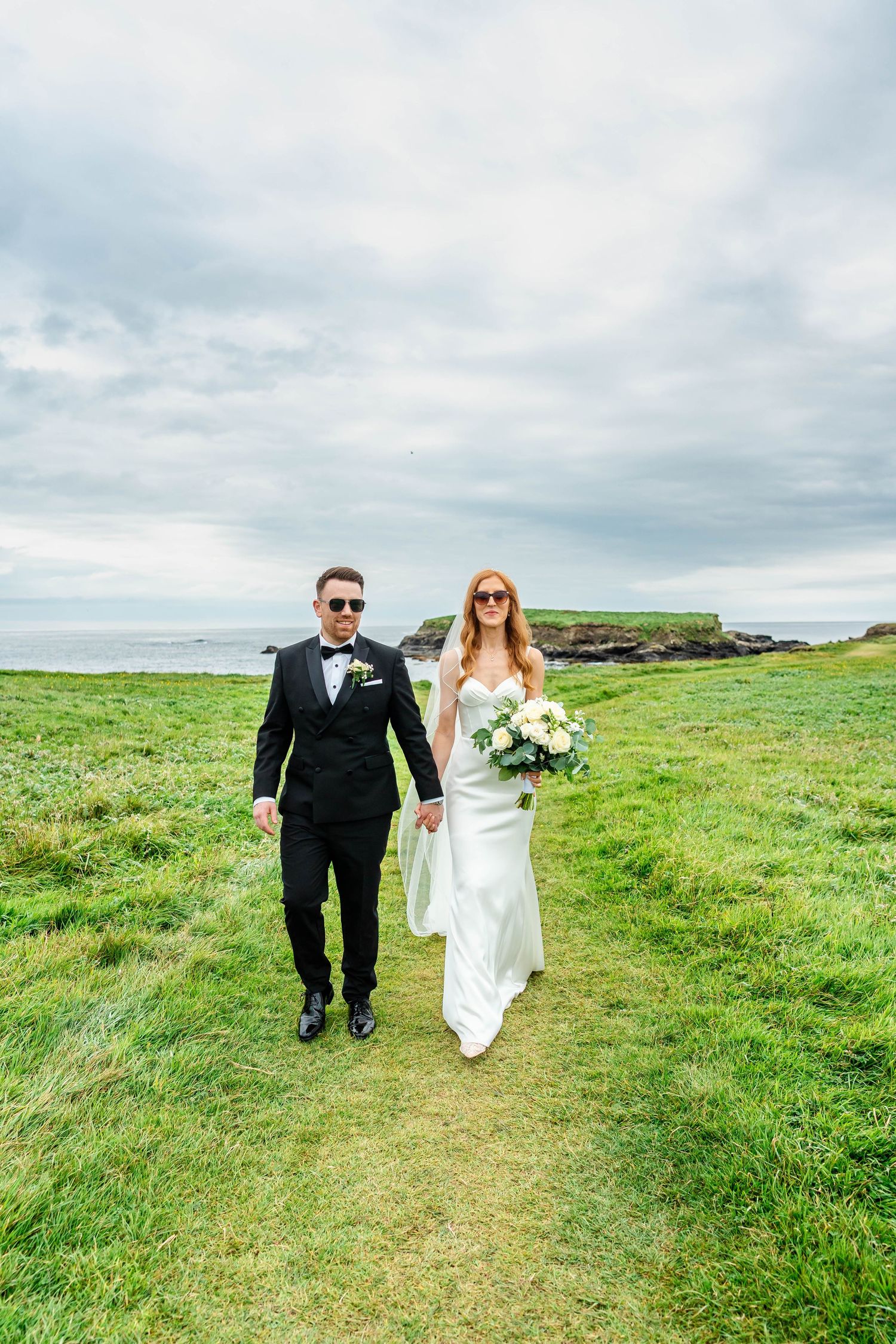 Bride and groom walking through windswept coastal grassland on their wedding day.