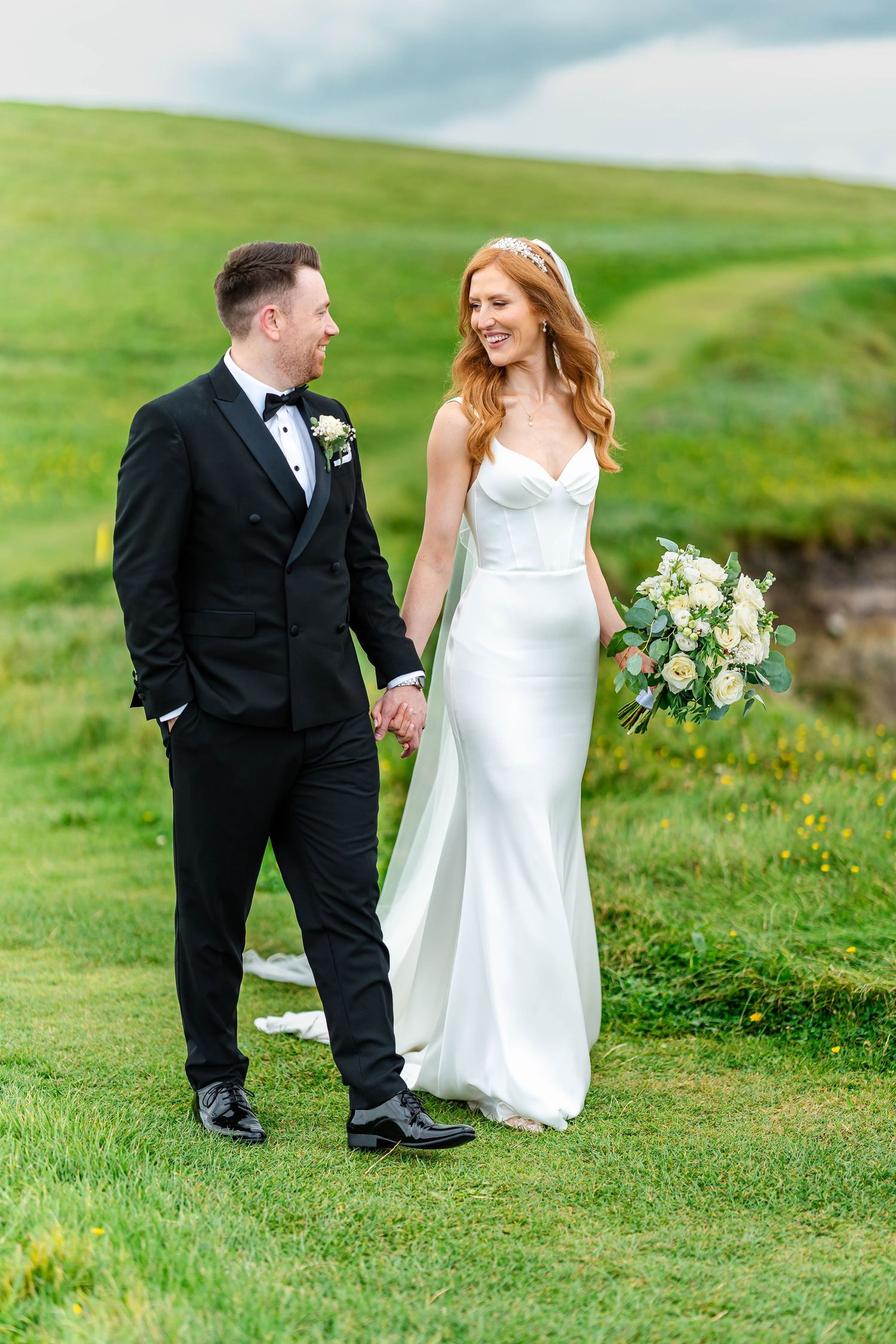 Couple walks hand in hand on lush green cliffside during their wedding.