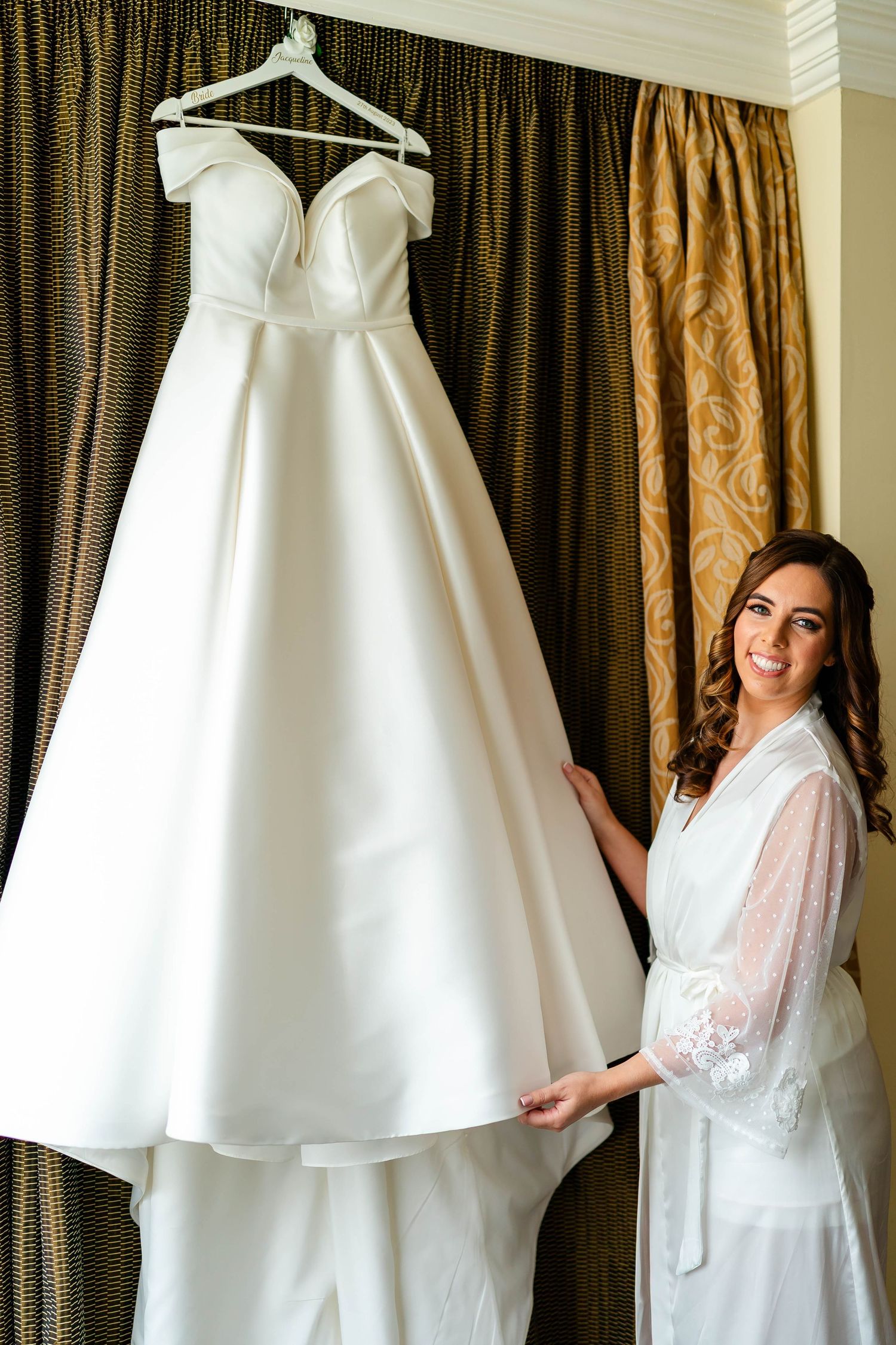 Elegant white wedding gown displayed against luxurious brown curtains.