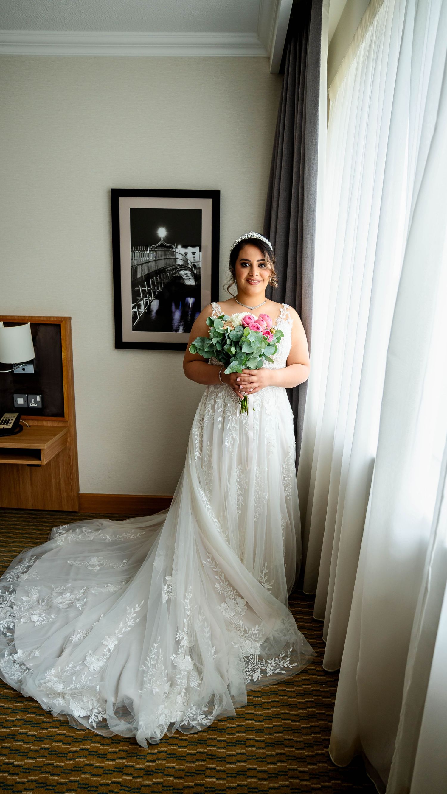 A beautiful lace wedding gown with a flowing train displayed in a hotel room setting.
