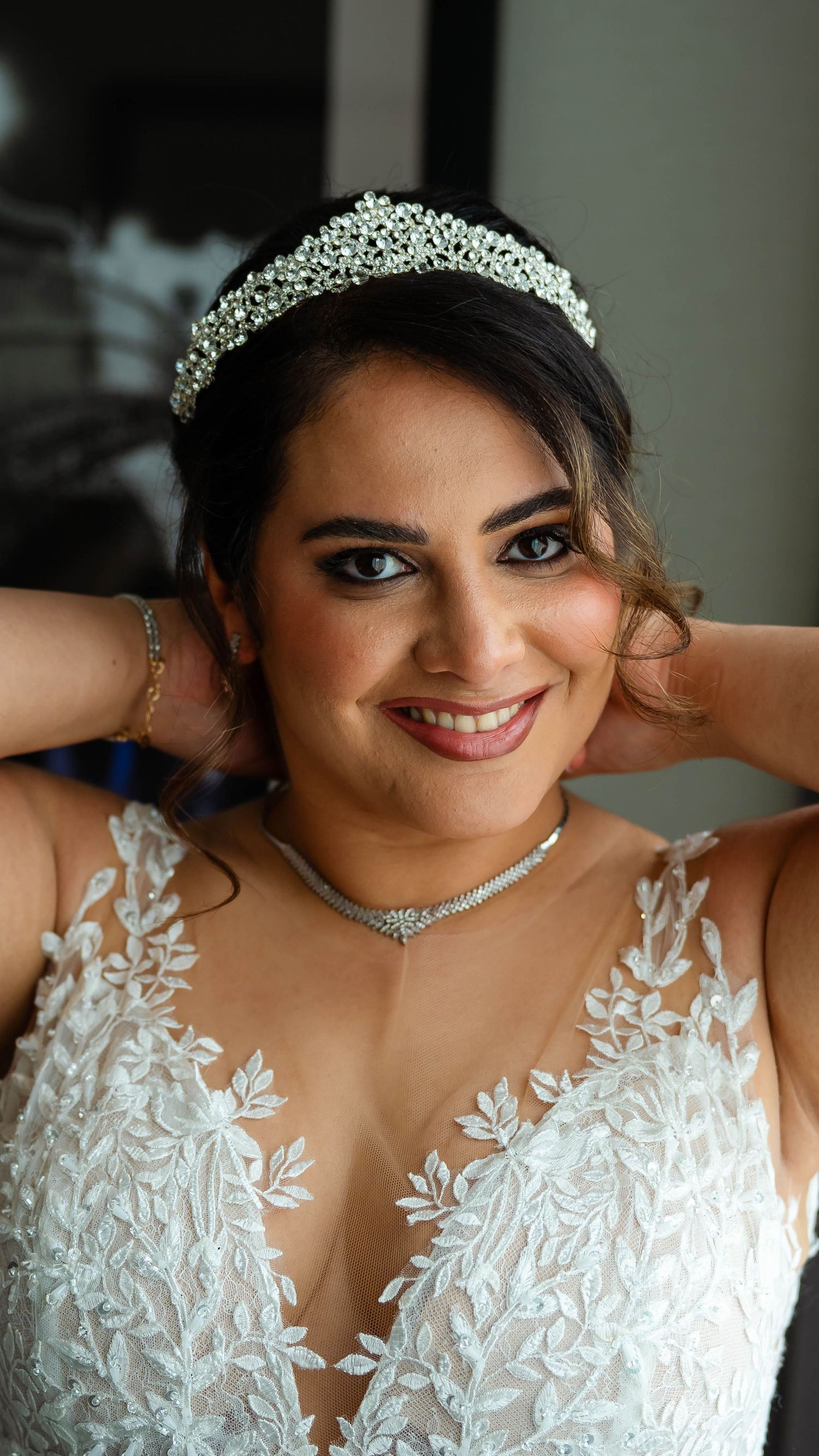 Smiling bride in a white lace wedding gown with intricate floral details and crystal tiara.