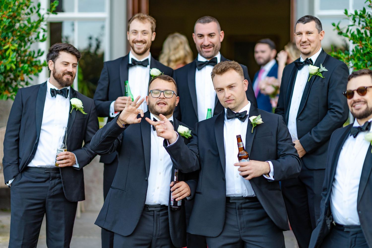 Groomsmen in black tuxedos and bowties share laughs and drinks during outdoor wedding festivities.