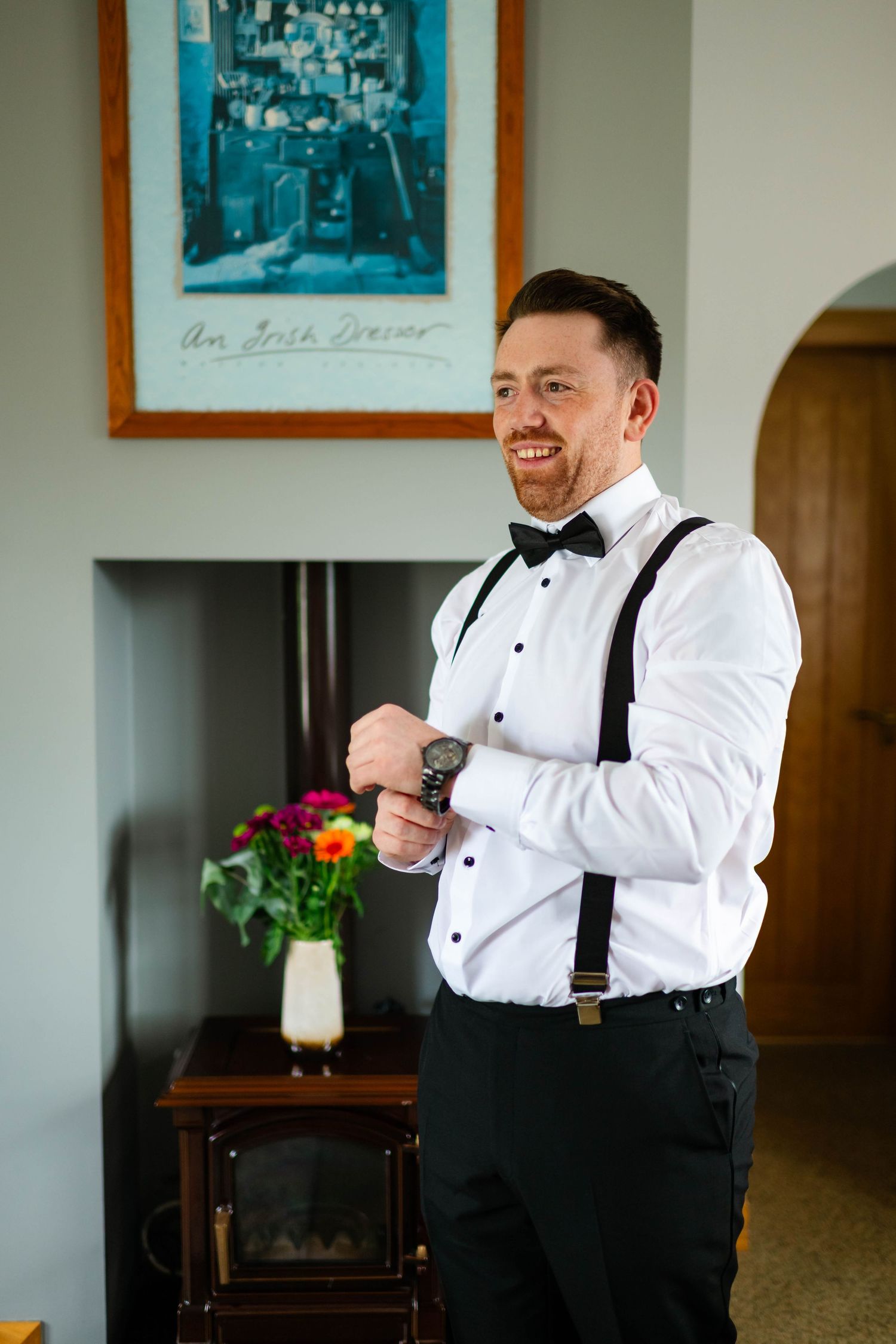Man adjusting suspenders while getting dressed in formal wear with framed artwork in background.