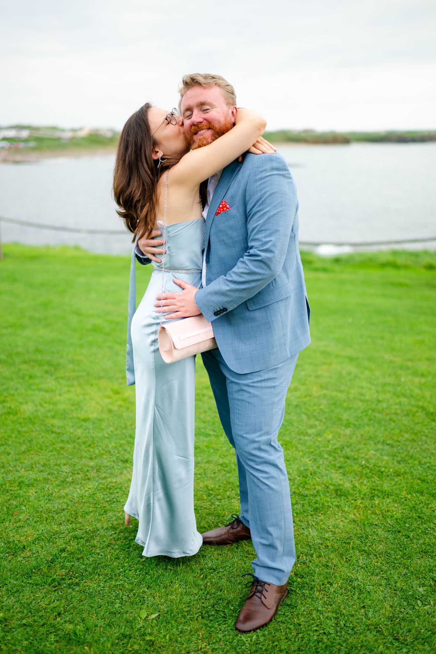 Couple embraces during intimate moment on grass overlooking waterfront.
