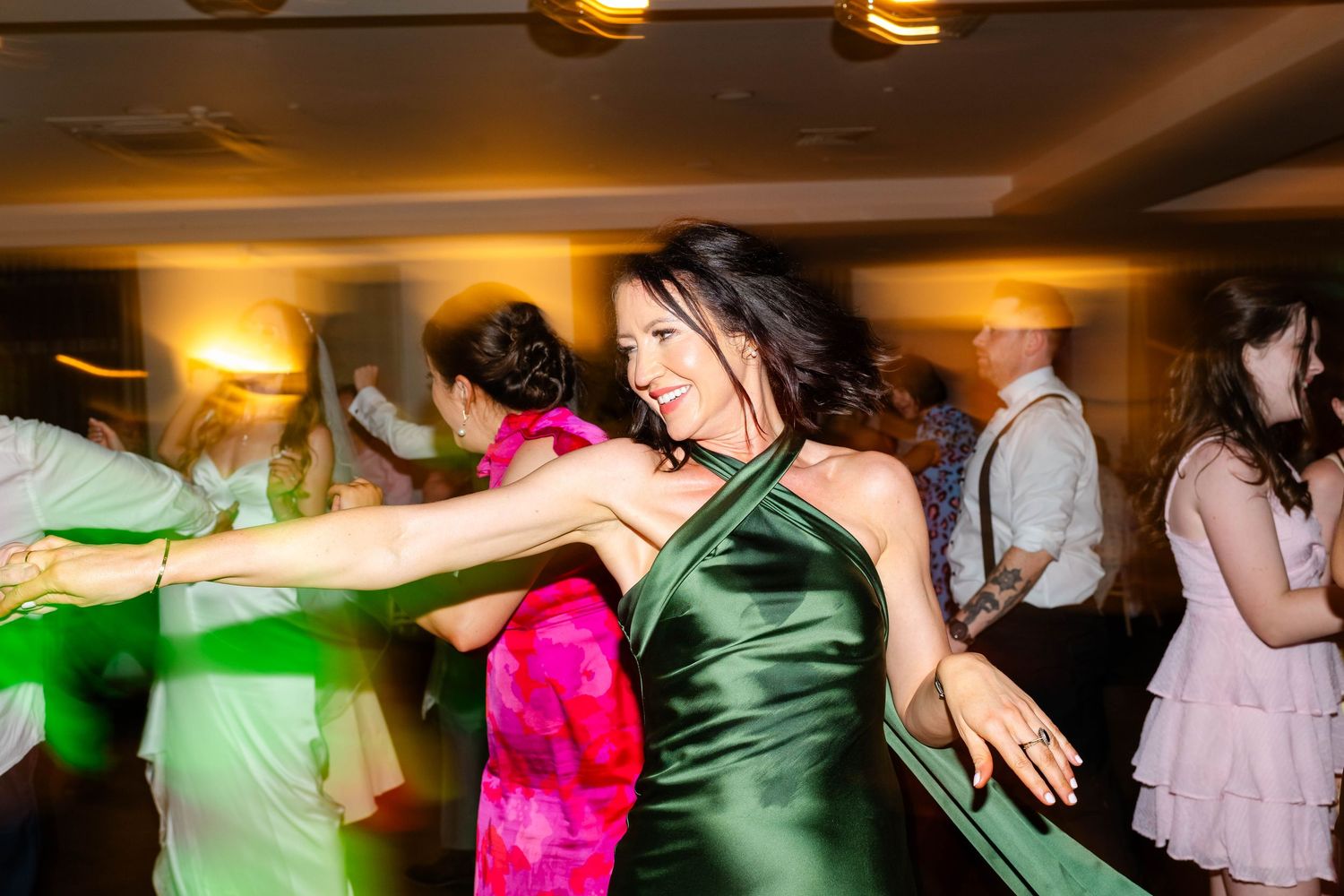Wedding party dancing in a line formation under warm ambient lighting at an indoor venue.