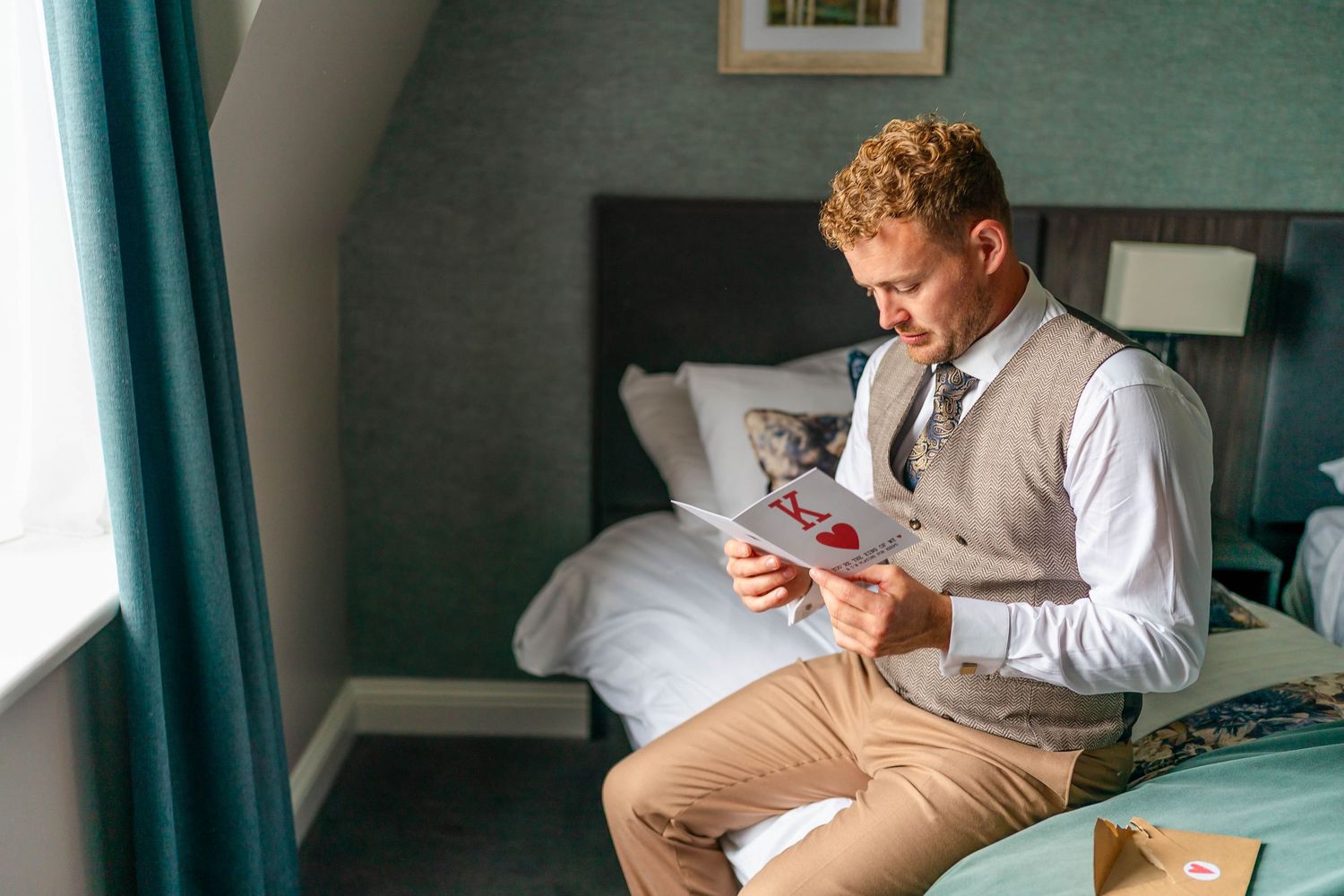 Person in white shirt and vest reads letter while sitting by window.